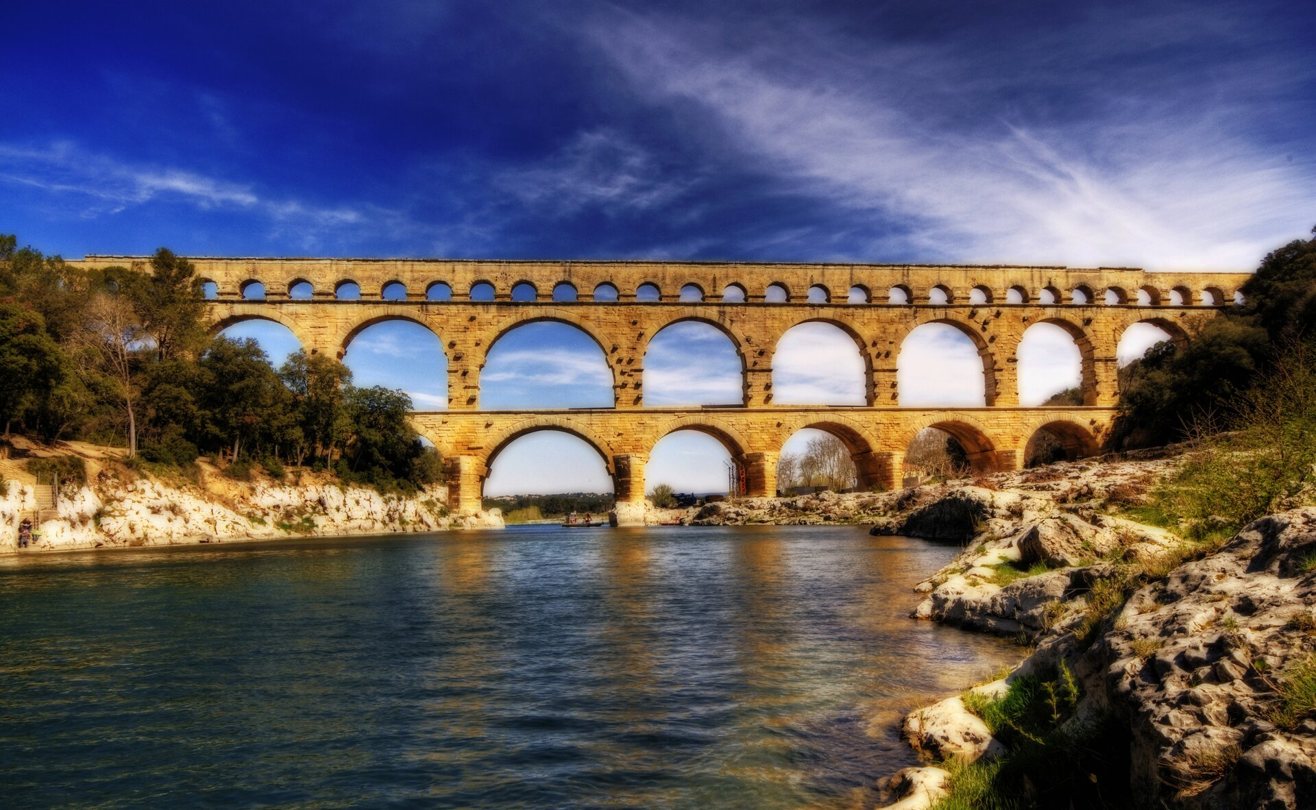 Ilustracja przedstawia Akwedukt Pont du Gard, ok. 26 p.n.e. - 19 p.n.e., obiekt z listy światowego dziedzictwa UNESCO we Francji. W kadrze, w tle znajduje się trójkondygnacyjny akwedukt. Na pierwszym planie znajduje się rzeka wraz z obydwoma brzegami i roślinnością po obu stronach zdjęcia, wykonanego podczas dnia, na tle lekko zachmurzonego, niebieskiego nieba. Przęsła akweduktu są szerokie, jednakowe na dwóch kondygnacjach. Kondygnacja najwyższa składa się z ciągu bardzo niskich arkad, pozostających w kontraście do dwóch pozostałych.
