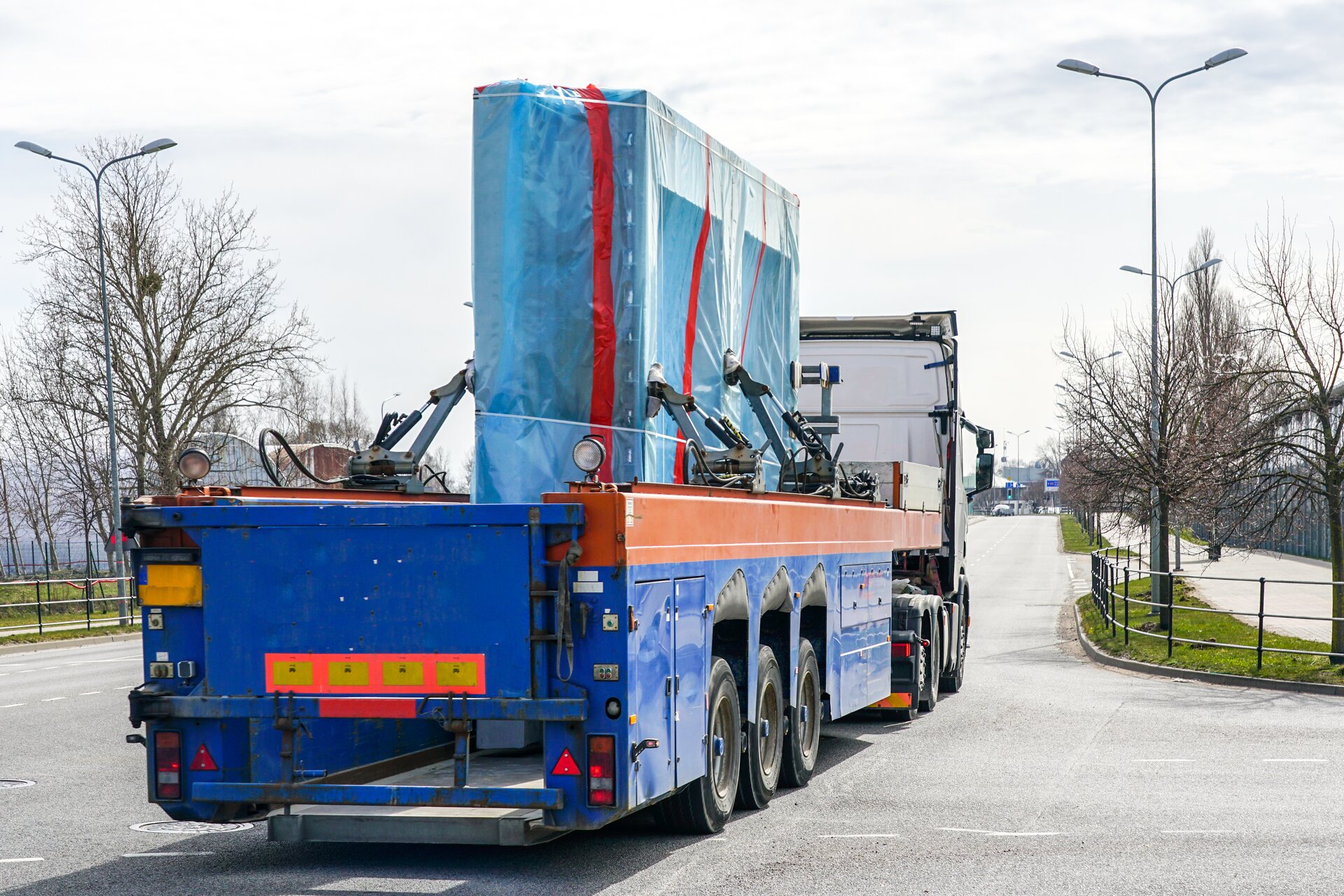 Zdjęcie przedstawia samochód transportowy jadący ulicą. Na platformie transportowej znajduje się prefabrykowana ściana betonowa. Ładunek zabezpieczony jest plandeką i wspornikami.
