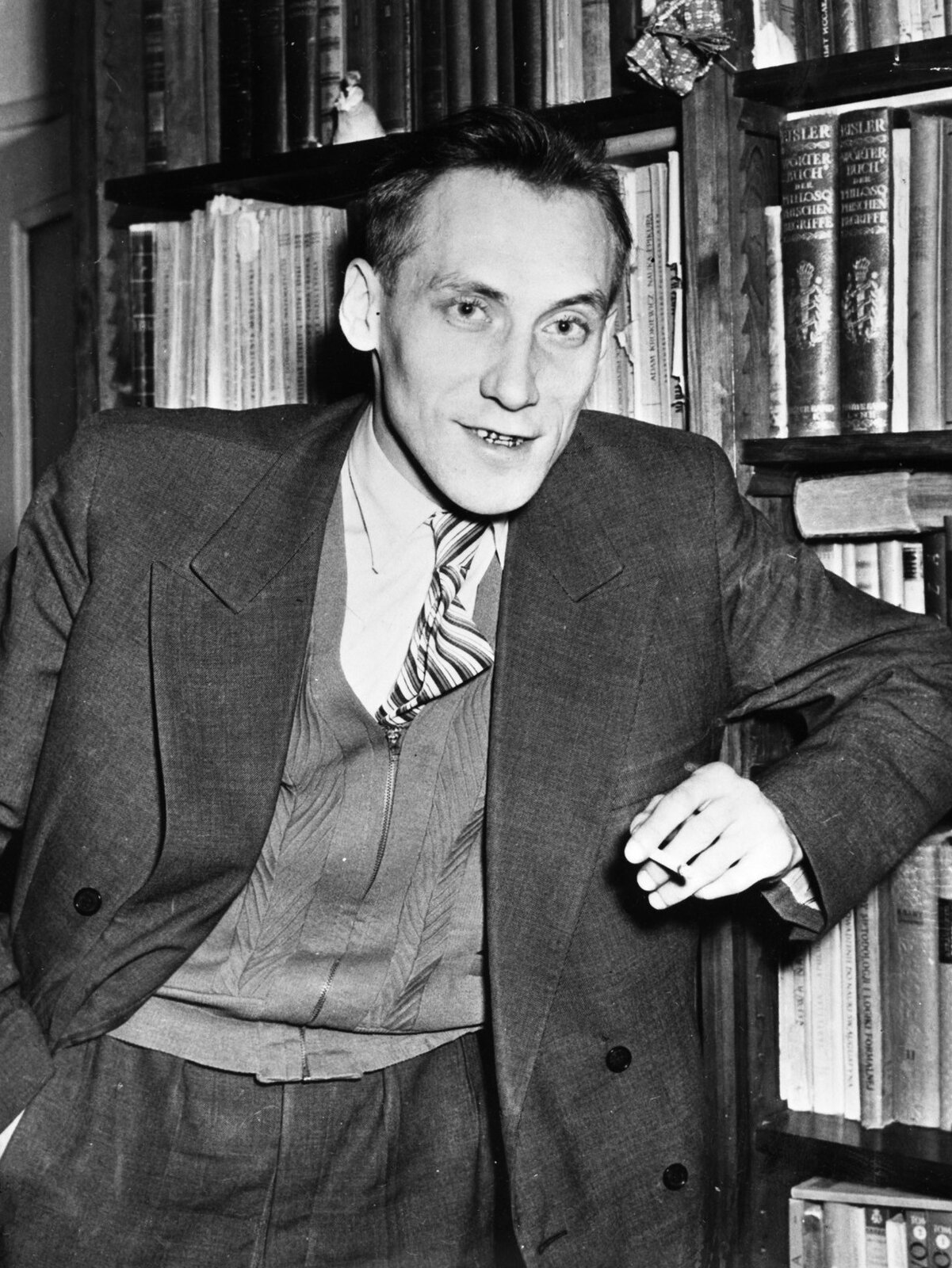 Zdjęcie przedstawia dojrzałego mężczyznę. Ma lekko zarysowane zmarszczki na pociągłej twarzy oraz krótkie włosy. Ubrany jest w garnitur. W ręku trzyma papieros. Za mężczyzną stoi półka z książkami.