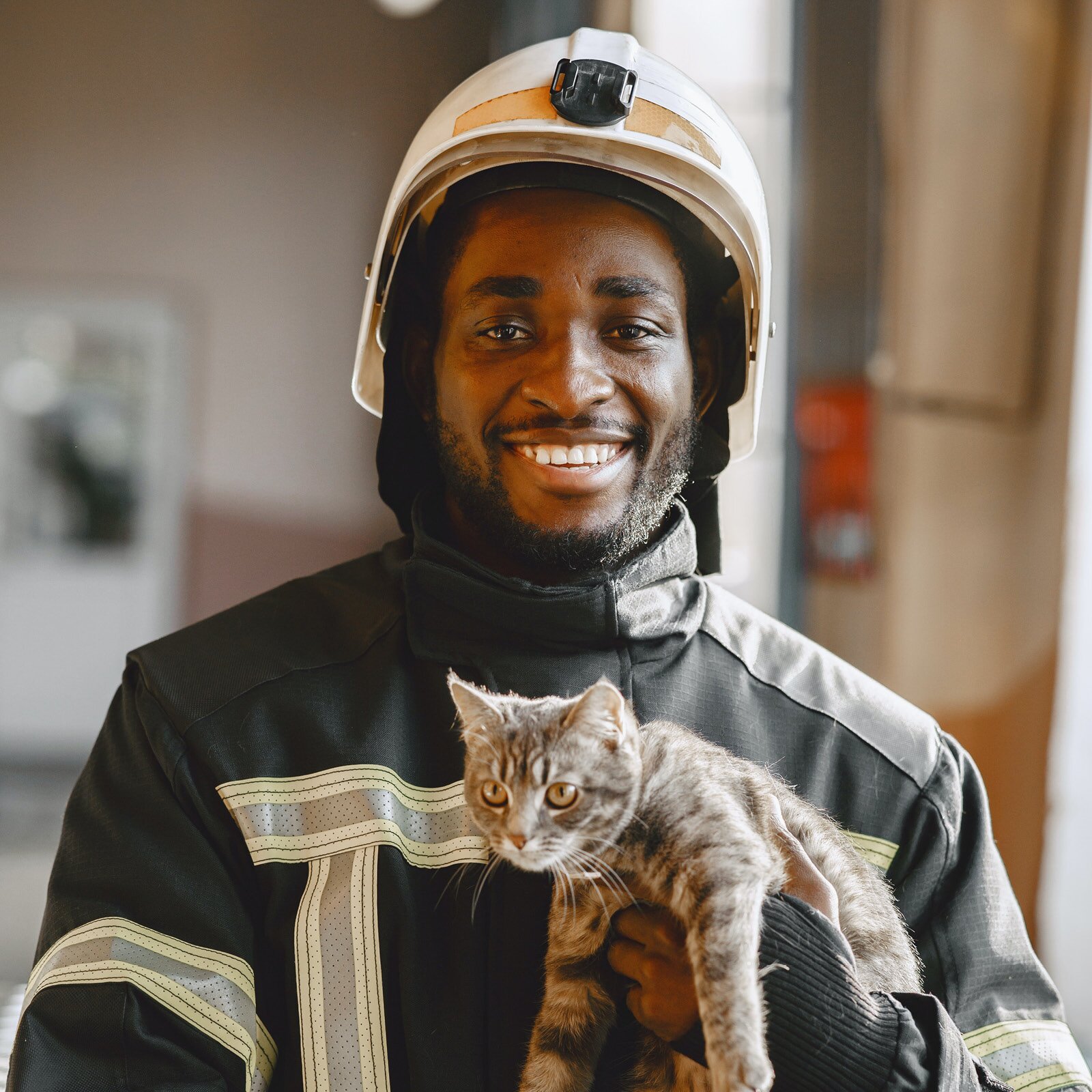 Ilustracja przedstawia uśmiechniętego strażaka, stojącego w remizie. Strażak ubrany jest w strój ochronny, złożony z białego hełmu i czarnego munduru z elementami odblaskowymi. Strażak trzyma w dłoni kota.