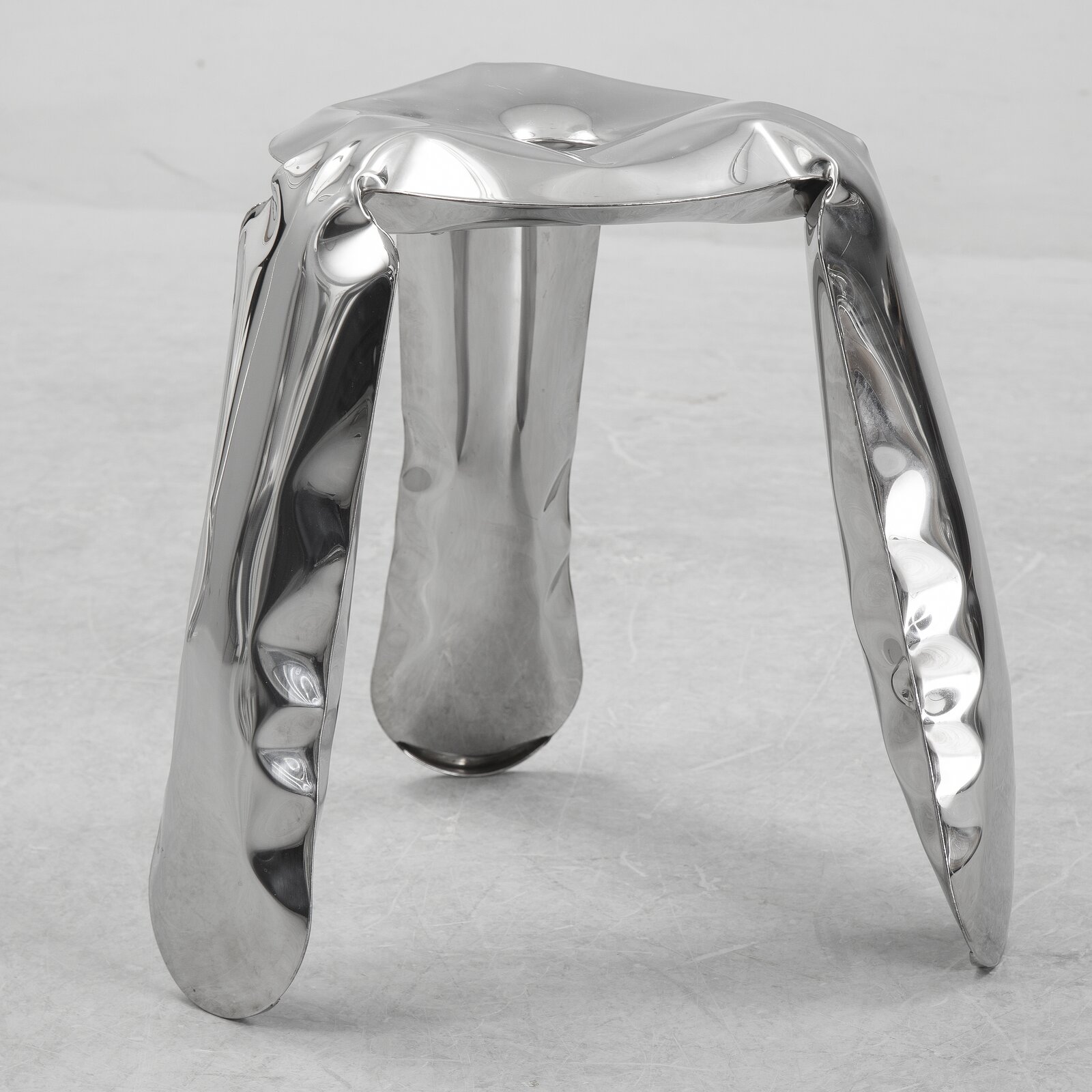 Zdjęcie przedstawia aluminiowy stołek. Jest on w srebrnym kolorze o błyszczącej fakturze, a od jego powierzchni odbija się otoczenie oraz światło. Materiał przypomina dmuchany balon, wydaje się miękki. Stołek posiada trójkątną część do siedzenia, z okrągłym wgnieceniem w środku i z trzech rogów trójkąta wychodzą trzy nogi, zaokrąglone przy ziemi. Zdjęcie wykonano na tle szarej podłogi.