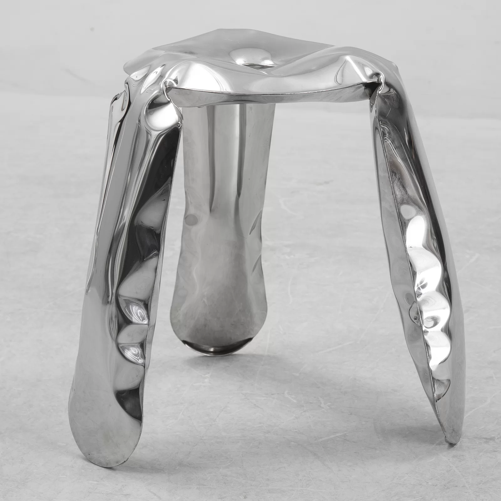 Zdjęcie przedstawia aluminiowy stołek. Jest on w srebrnym kolorze o błyszczącej fakturze, a od jego powierzchni odbija się otoczenie oraz światło. Materiał przypomina dmuchany balon, wydaje się miękki. Stołek posiada trójkątną część do siedzenia, z okrągłym wgnieceniem w środku i z trzech rogów trójkąta wychodzą trzy nogi, zaokrąglone przy ziemi. Zdjęcie wykonano na tle szarej podłogi.