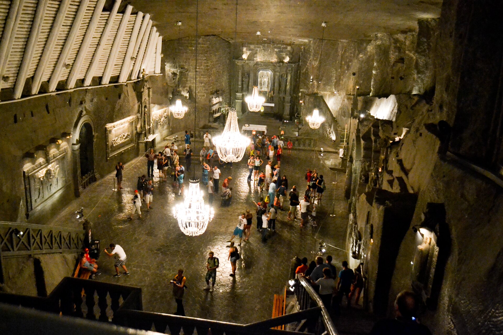 Zdjęcie ukazuje obszerną komnatę w kopalni soli. To kaplica świętej Kingi, w której znajdują się relikwie świętej. Z sufitu zwisają okazałe żyrandole. Podłoga połyskuje. Na ścianach znajdują się płaskorzeźby. Do kaplicy prowadzą schody. W pomieszczeniu jest sporo odwiedzających.