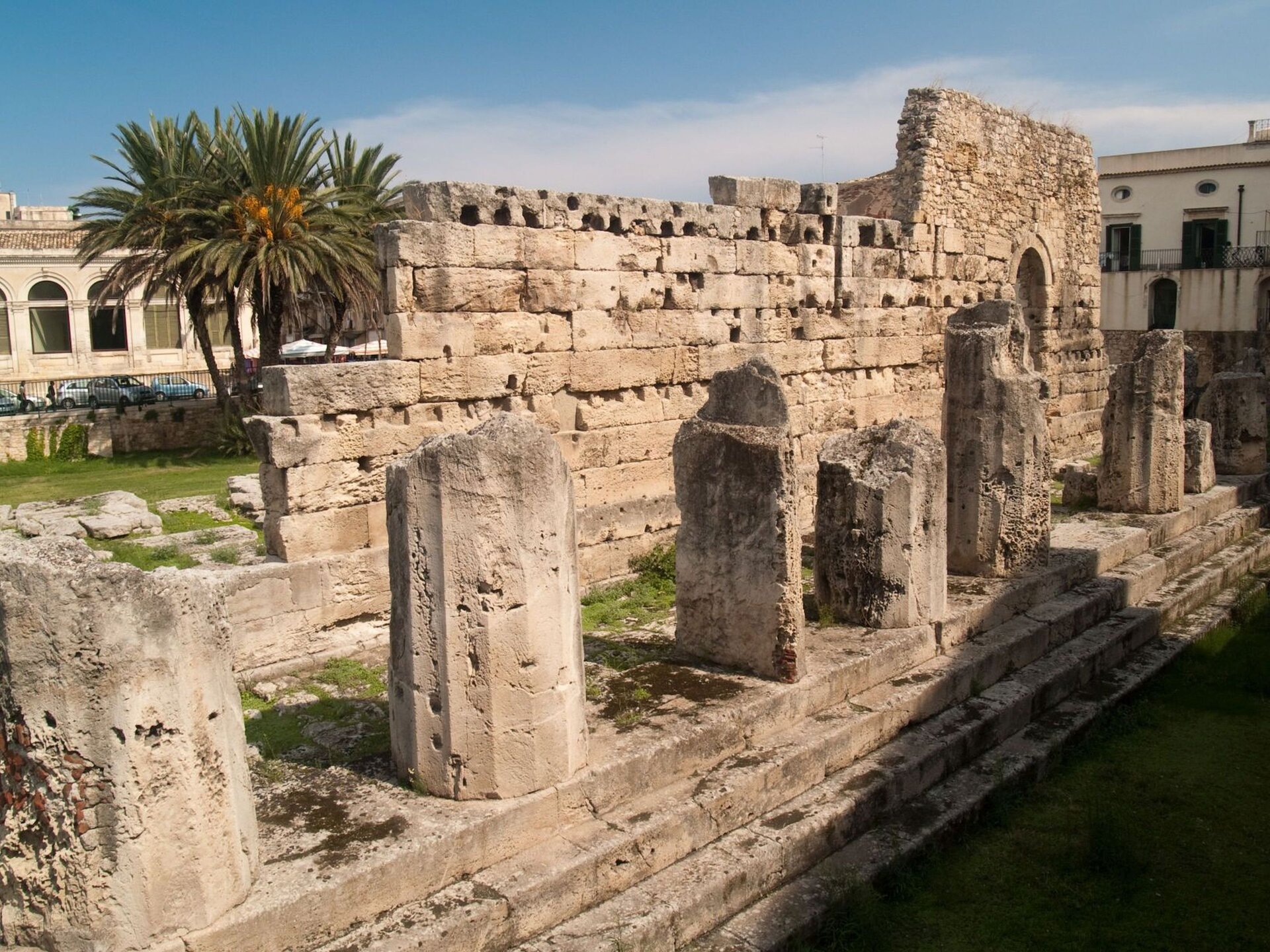 Ilustracja przedstawia Ruiny Świątyni Apolla na Wyspie Ortygia w Syrakuzach. Na zdjęciu widoczne są fragmenty podstawy i fundamentów doryckiej świątyni. Pozostałości po świątyni wskazują, iż była olbrzymia i otaczały ją masywne kolumny. W tle widoczna jest miejska zabudowa oraz palmy.