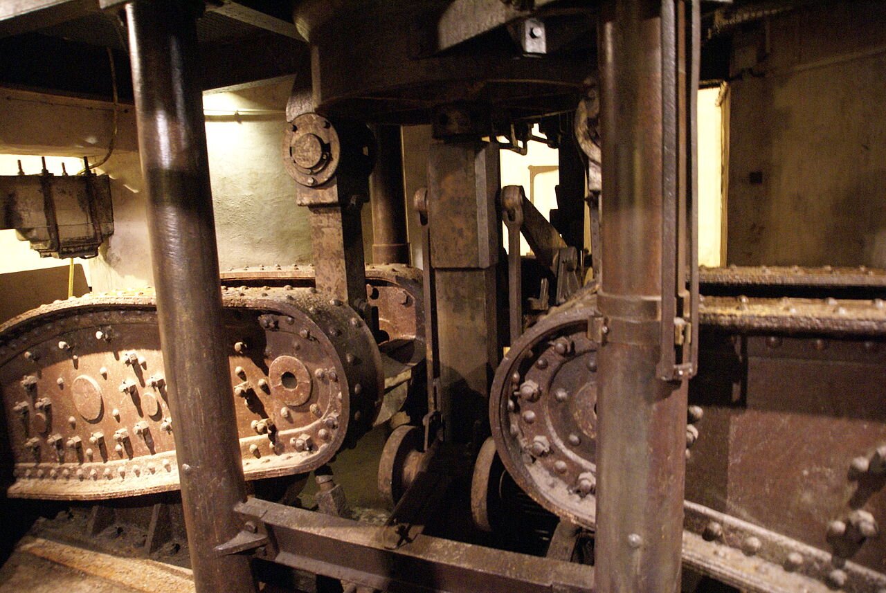 Zdjęcie przedstawia działko z bronią. Po obu stronach są metalowe taśmy z nitami. Pomiędzy nimi są szyny na rolkach. Wszystkie elementy są metalowe.