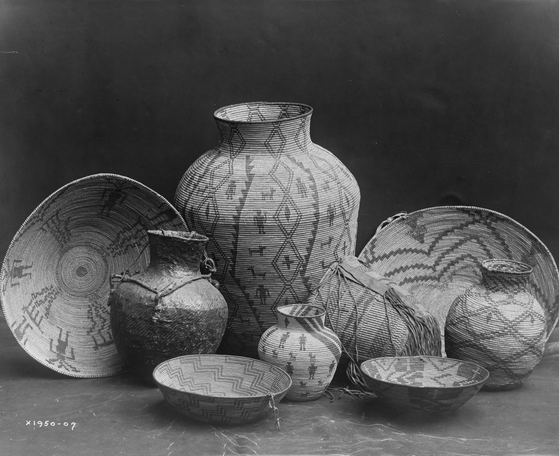 Czarno‑biała fotografia przedstawia wyplatane z traw przedmioty codziennego użytku: dzbany, miski i kosz. Zostały ozdobione etnicznymi wzorami Apaczów, Indian Ameryki Północnej. Na środku stoi duży dzban, ozdobiony motywami roślinnymi i zwierzęcymi, po jego bokach ustawiono dwie duże misy, przed nimi stoją trzy mniejsze dzbanki i dwie mniejsze miski oraz przewrócony kosz. Jeden z mniejszych dzbanków powleczony jest wypaloną gliną, błyszczy się i nie ma na nim wzorów.