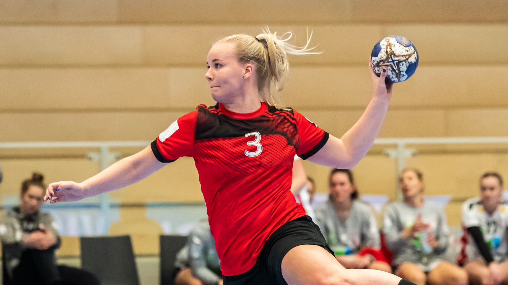 Zdjęcie przedstawia kobietę w sportowej koszulce rzucającą piłką ręczną. W tle widoczne są  kobiety siedzące na ławce na tle pustych trybun.