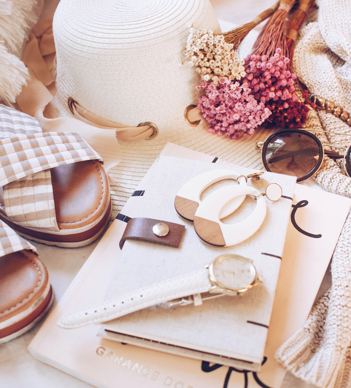Zdjęcie przedstawia różne dodatki do ubrania, które leżą koło siebie. Wszystkie dodatki są w jasnych kolorach i pasują do siebie kolorystycznie. Widoczne są sandały w biało‑beżową kratkę, kolczyki w kształcie elipsy, biały zegarek na rękę, okulary przeciwsłoneczne, biały, słomkowy kapelusz, korale i kremowy szalik. Zegarek i kolczyki leżą na dwóch notatnikach, większym i mniejszym, również pasujących kolorystycznie do reszty. Na górze zdjęcia znajduje się pęk suszonych kwiatów.