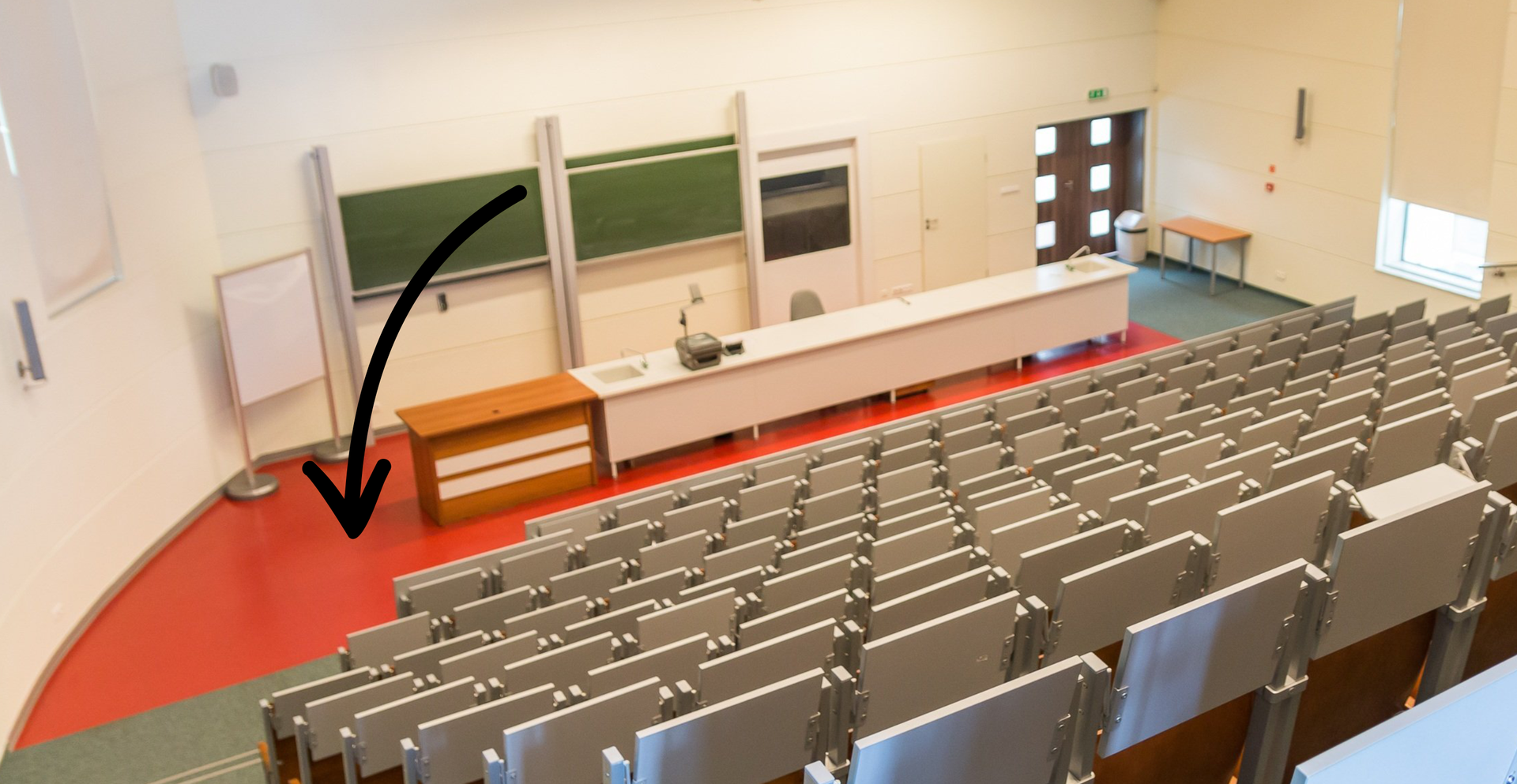 Zdjęcie przedstawia aulę. Widoczne są rzędy krzeseł. W centralnym miejscu znajduje się scena. Na scenie znajduje się duże biurko i tablice szkolne. Strzałka na ilustracji skierowana jest na scenę.