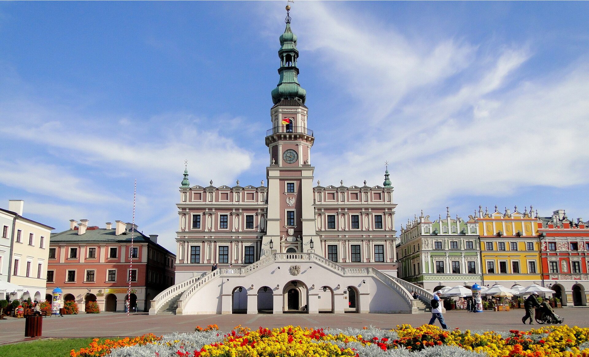 Fotografia barwna prezentująca frontową elewację ratusza w Zamościu. To trzypoziomowy budynek z sześciopiętrową wieżą, na której znajduje się zegar z 1770 r. Wieża zwieńczona jest cebulastym daszkiem, zwanym dachem hełmowym lub po prostu hełmem. Do wejścia ratusza, usytuowanym na drugim poziomie budowli, prowadzą z dwóch stron  symetryczne, półkoliste, wachlarzowe schody, wsparte na pięciu arkadach, czyli kolumnach połączonych ze sobą łukiem.  Color photography presenting the front elevation of the town hall in Zamość. It is a three-storey building with a six-storey tower, on which there is a clock from 1770. The tower is topped with an onion roof, called a helmeted roof or simply a helmet. To enter the town hall, located on the second level of the building, they lead from two sides symmetrical, semicircular, fan-shaped stairs, supported on five arcades, ie columns connected with each other by an arch.