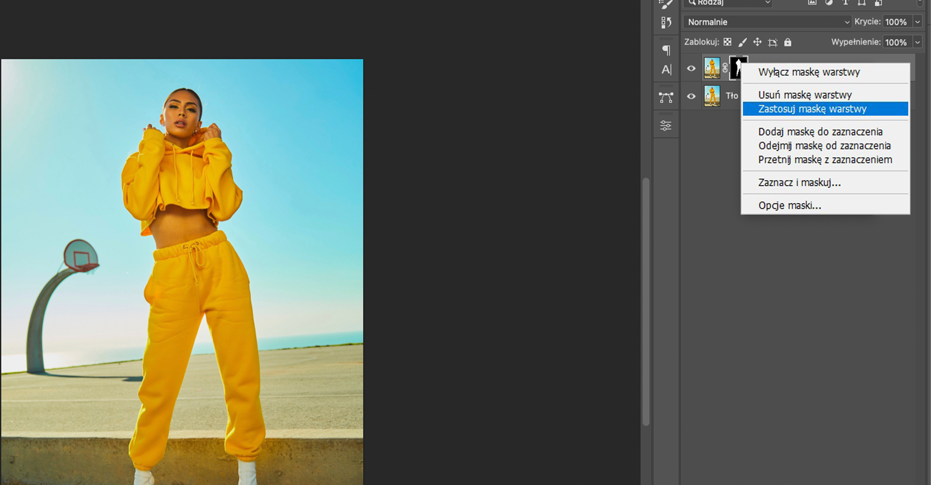 Ilustracja przedstawia okno programu. W obszarze roboczym jest modelka ubrana w dresowy komplet. W tle jest boisko do koszykówki. Zdjęcie zajmuje tylko lewą część obszaru roboczego. Po prawej stronie okna jest panel dotyczący Warstw. Tam rozwinięta lista. Zaznaczono opcję: Zastosuj maskę warstwę.  