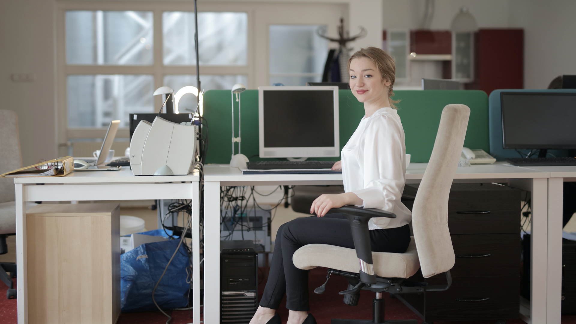 Zdjęcie przedstawia kobietę, która siedzi wyprostowana w fotelu przy biurku. Na biurku znajduje się monitor komputerowy oraz drukarka.