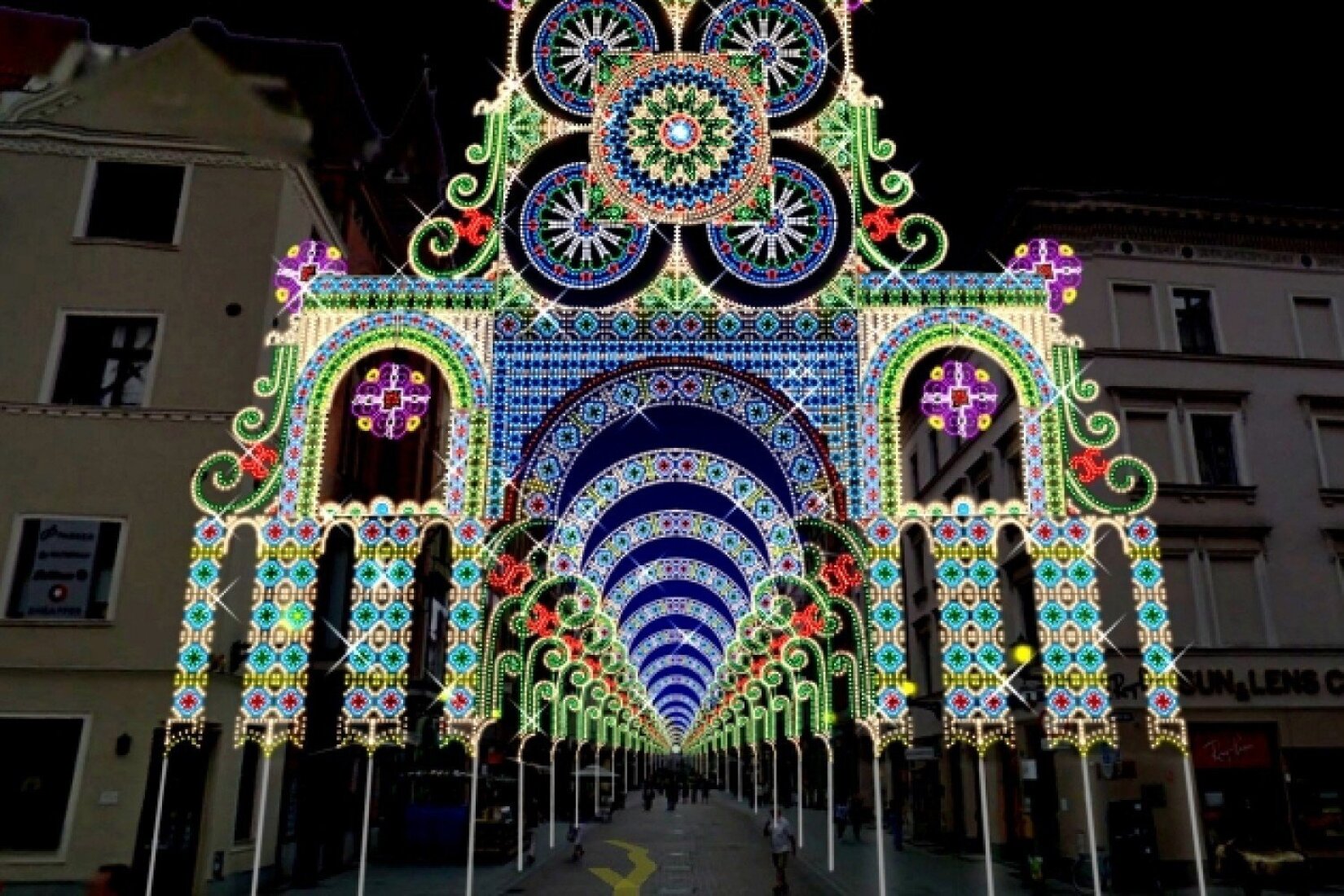 Ukazana zostaje fotografia przedstawiająca przejście pomiędzy dwoma beżowymi budynkami. Przejście oświetlone jest wieloma kolorowymi światłami. Światła składają się na spójną całość tworząc korytarz świetlny imitujący świetliste niebo.