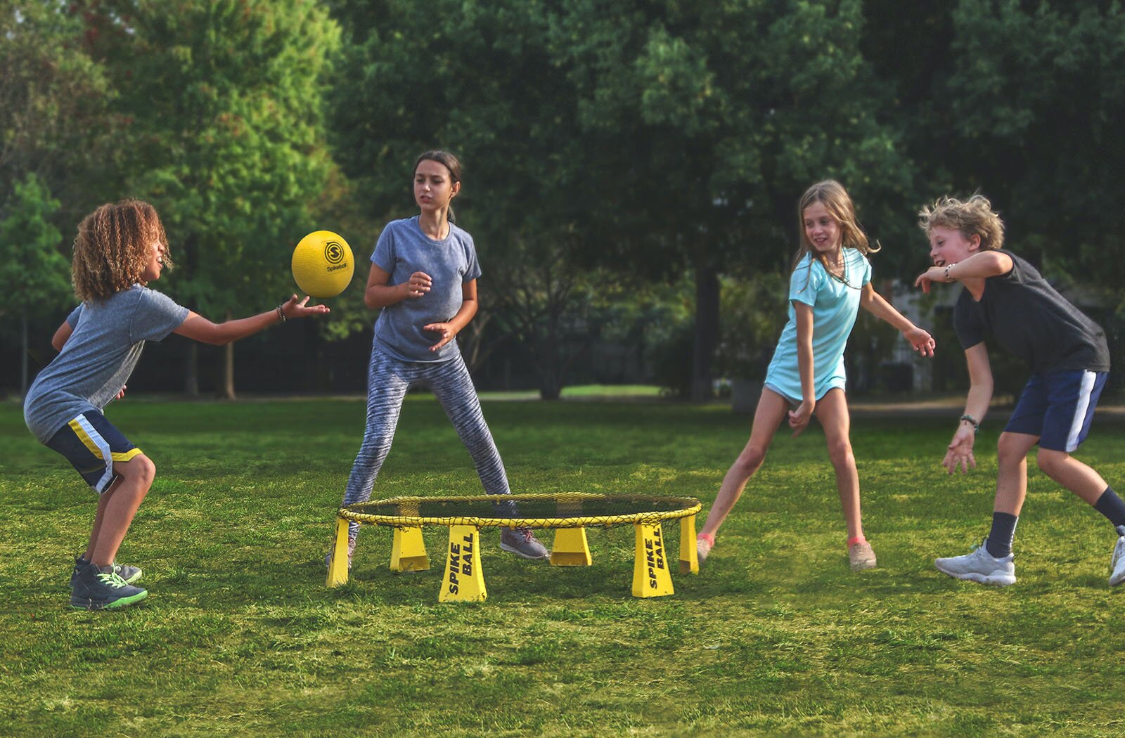 Zdjęcie przedstawia czwórkę dzieci (dwie dziewczynki i dwóch chłopców) w wieku około 12 lat grających w plenerze w grę spike ball za pomocą żółtej piłki. Dzieci ubrane są w sportowe stroje: koszulki z krótkim rękawem, szorty lub leginsy. Na trawie pomiędzy nimi stoi mała, żółta trampolina, na której nogach widoczny jest napis w języku angielskim: SPIKE BALL. W tle widoczne są drzewa.