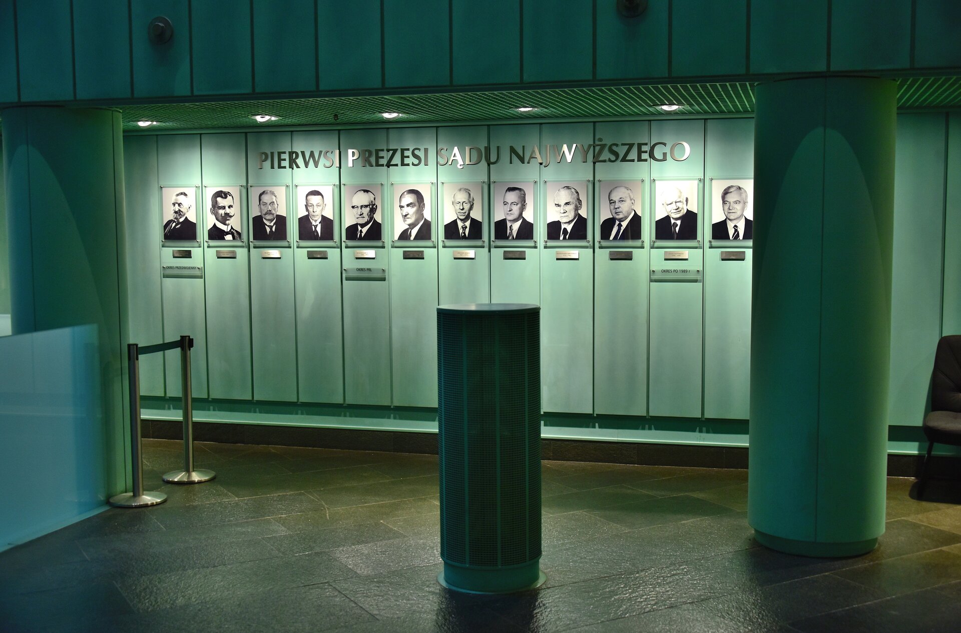 Zdjęcie przedstawia pomieszczenie. Na ścianie wiszą zdjęcia mężczyzn w garniturach. Pod każdym ze zdjęć znajduje się tabliczka z podpisem. Nad zdjęciami jest napis Pierwsi Prezesi Sądu Najwyższego.