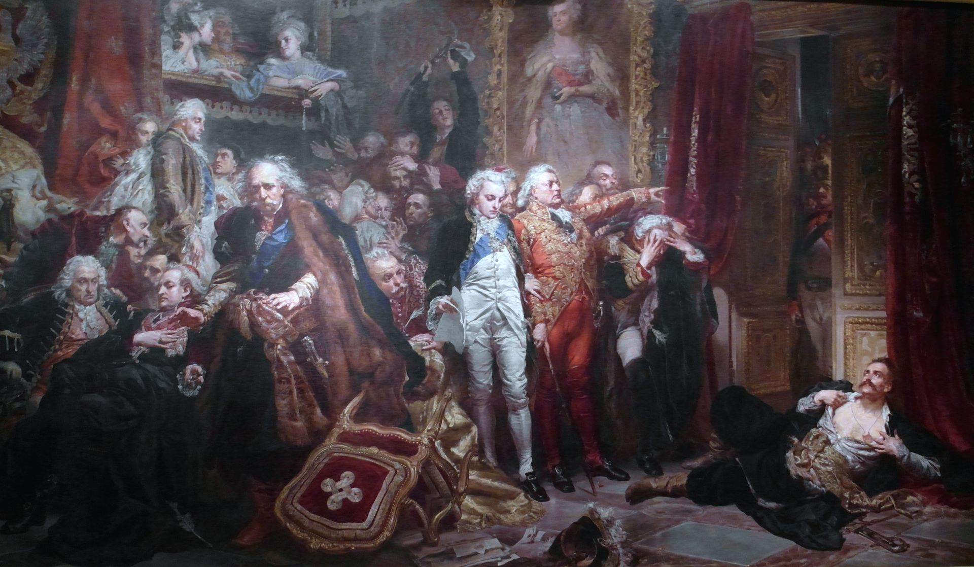 Ilustracja przedstawia obraz Jana Matejki pod tytułem "Rejtan - upadek Polski".  Przedstawia on mężczyzn zgromadzonych w królewskiej sali. Wszyscy są ubrani w odświętne szaty, wyszywane złotymi nićmi.  Na głowach mają peruki, a przy bokach szable. W rogu obrazu, pod ścianą leży mężczyzna ukazujący swoją pierś. W dramatycznym geście rozrywa on swoją koszulę. Jeden z mężczyzn wskazuje mu wyjście. Na obrazie panuje ogólny chaos, krzesło jest przewrócone, na twarzach ludzi malują się różne emocje.