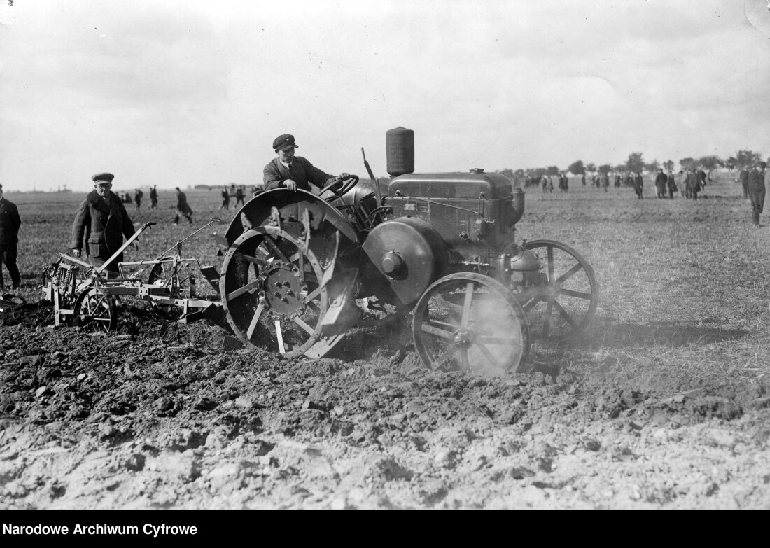 Na czarno – białej fotografii przedstawione są prace polowe przy użyciu ciągnika mechanicznego w Wielkopolsce w 1928 roku. Na pierwszym planie widoczny jest traktorzysta siedzący na ciągniku. Na głowie ma ciemną czapkę z daszkiem. Maszyna jest w ruchu – jej tylne koło jest sporo większe niż przednie, ma zaostrzone krawędzie i jest okryte błotnikiem. Do traktora przyczepiona jest maszyna rolnicza. Za ciągnikiem idzie drugi mężczyzna, na dalszym planie znajdują się liczni ludzie.