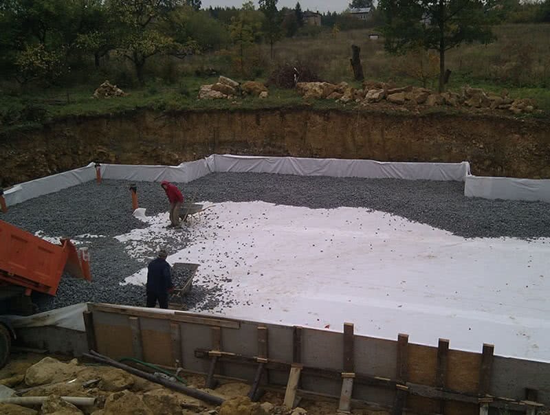 Zdjęcie przedstawia rozkładanie szkła piankowego. Pracownicy rozwożą je taczkami po obszarze oznaczonym białą folią. Samo szkło piankowe jest drobne, ma kulisty kształt i czarny kolor.