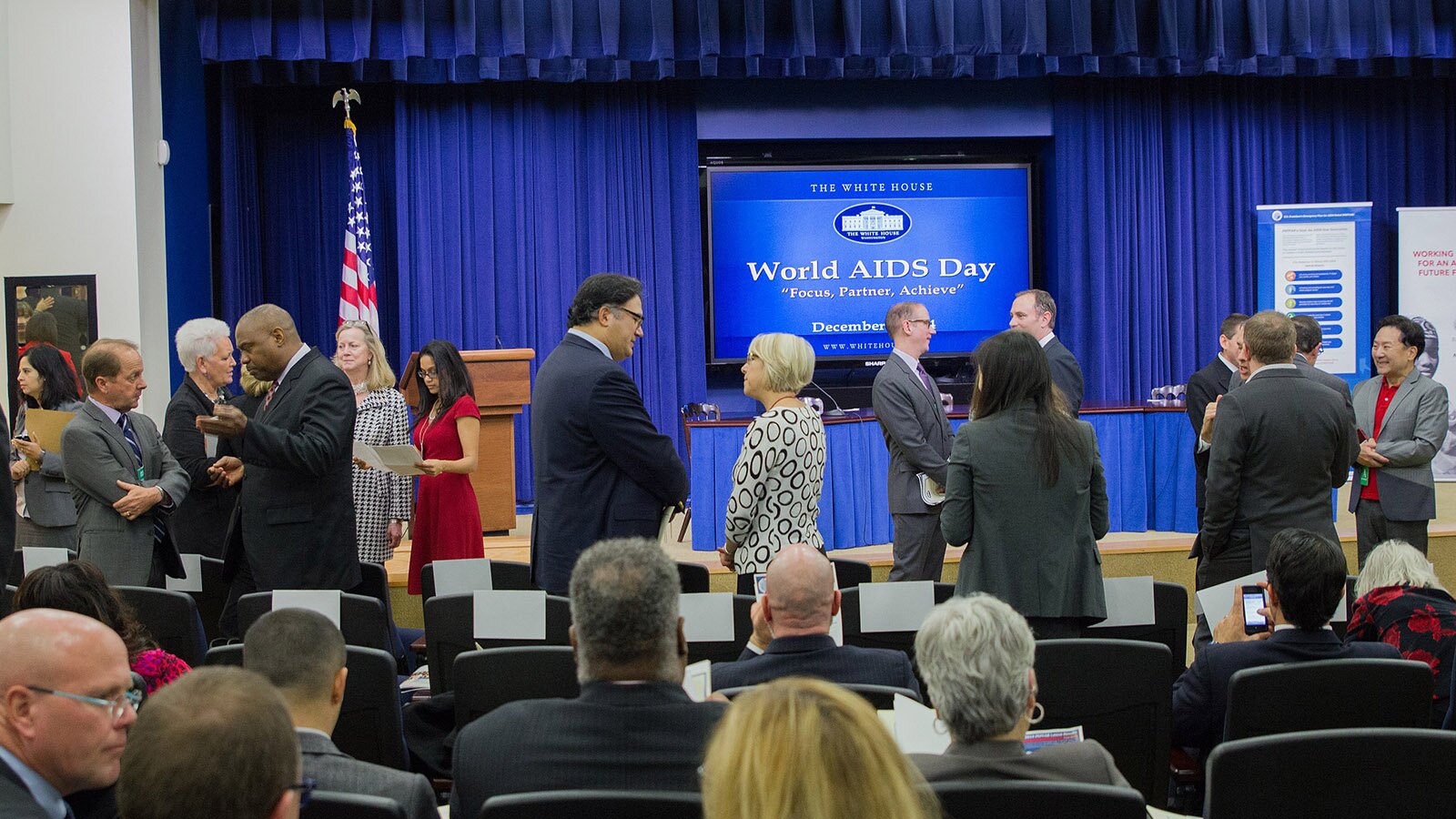 Na zdjęciu widoczne osoby biorące udział w konferencji. Na ekranie telebimu wyświetlono napis: WORLD AIDS DAY. 
