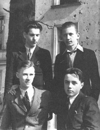 Zdjęcie przedstawia czterech młodych mężczyzn w garniturach. Dwóch z nich siedzi, dwóch pozostałych stoi za ich plecami. W tle drzewo i kamienica.