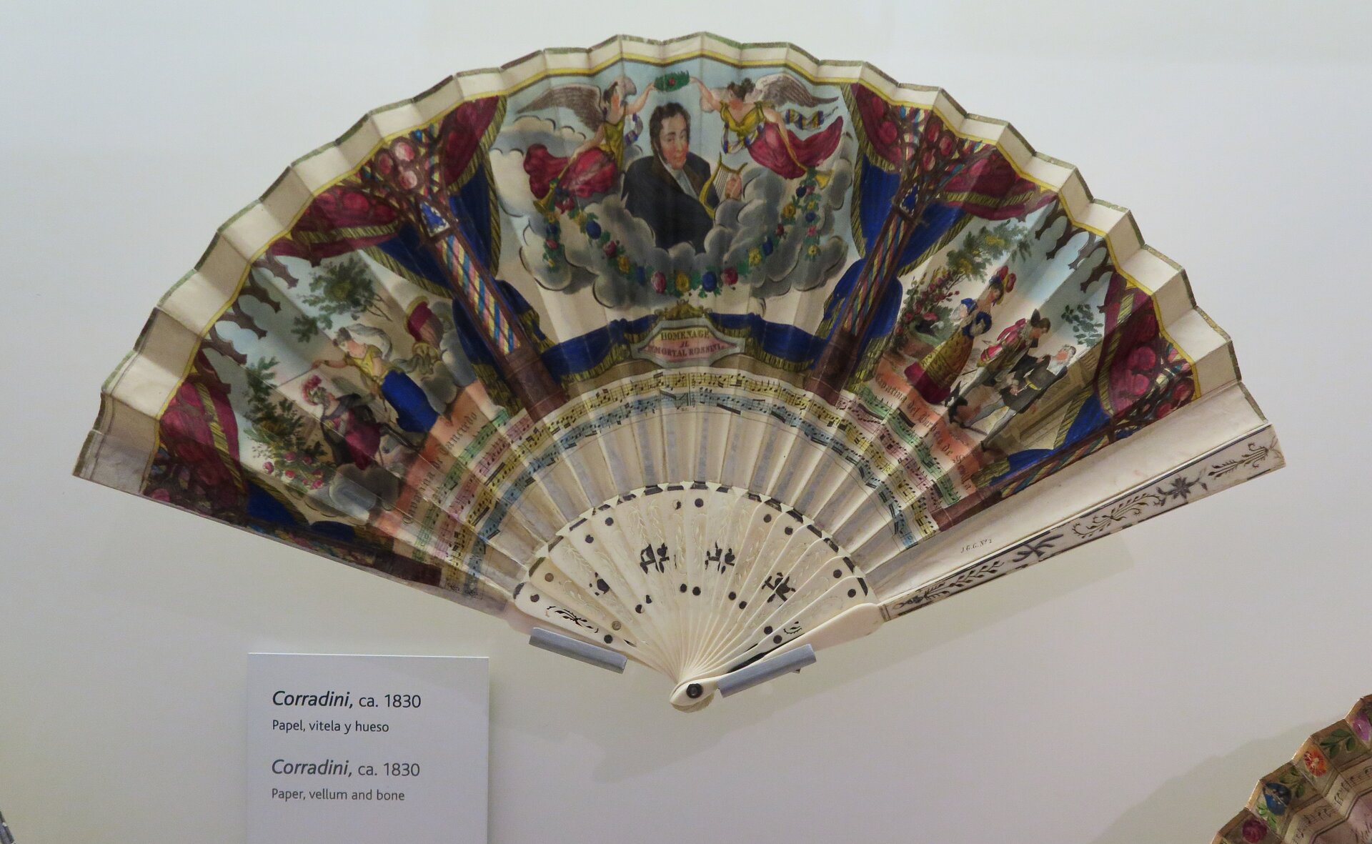 Kliknij, aby powiększyć. Zorientowana poziomo fotografia przedstawiająca ręcznie malowany wachlarz. Malunek pokrywa większą część jasnego materiału, z którego wykonano wachlarz. Składają się nań trzy sceny ujęte w ramy z draperii rozpiętych na kolumnach. Scena środkowa ukazuje popiersie mężczyzny w czarnym surducie wyłaniające się ze skłębionych obłoków. Trzyma on w dłoniach lirę grecką. Po bokach ponad obłokami dwie postaci uskrzydlonych kobiet w czerwonych sukniach. W jednej z rąk unoszą nad głową mężczyzny zielony wieniec, a w drugiej opadającą łukiem na obłoki girlandę kwiatową. Scena z lewej przedstawia kobietę unoszącą różdżkę nad rzymskim legionistą. Z prawej dama w długiej sukni i z kokardą upiętą na głowie, a obok zajęci rozmową mężczyźni. Bliższy kobiety ze szpadą u pasa w stroju okresu baroku, a drugi w renesansowym. U dołu wachlarza z lewej nieduża kartka z informacją o eksponacie.