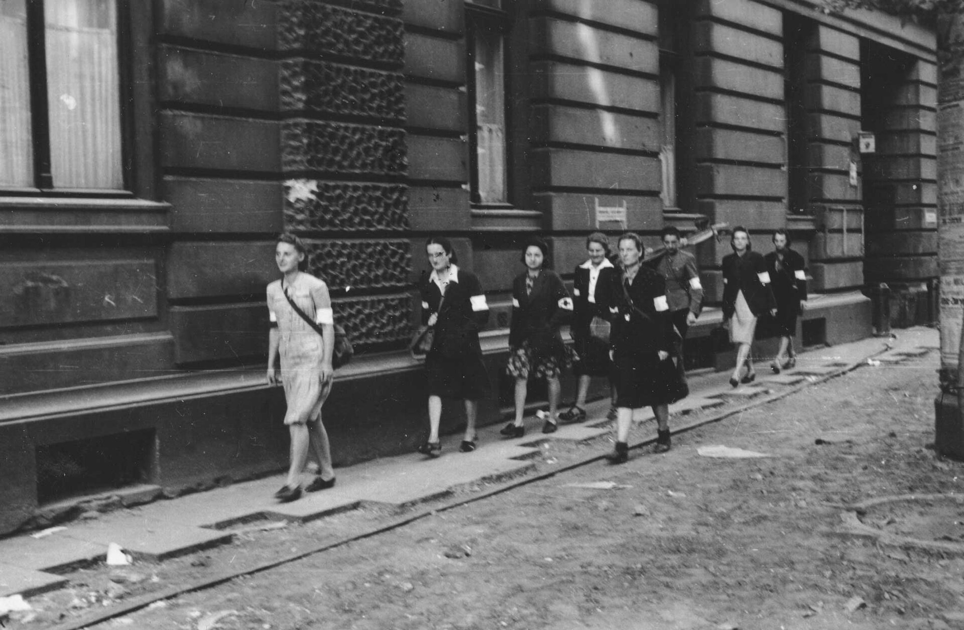 Zdjęcie przedstawia grupę sanitariuszek podczas powstania warszawskiego. To młode kobiety z przepaskami na dłoniach. Idą chodnikiem wzdłuż kamienicy.