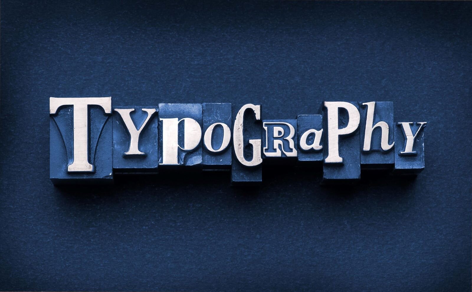 Zdjęcie przedstawia napis " Typography  [czytaj: typografi]". Na granatowym tle ułożono czcionki drukarskie o różnej wielkości i rodzaju. Litery wyróżniają się, gdyż są białe.