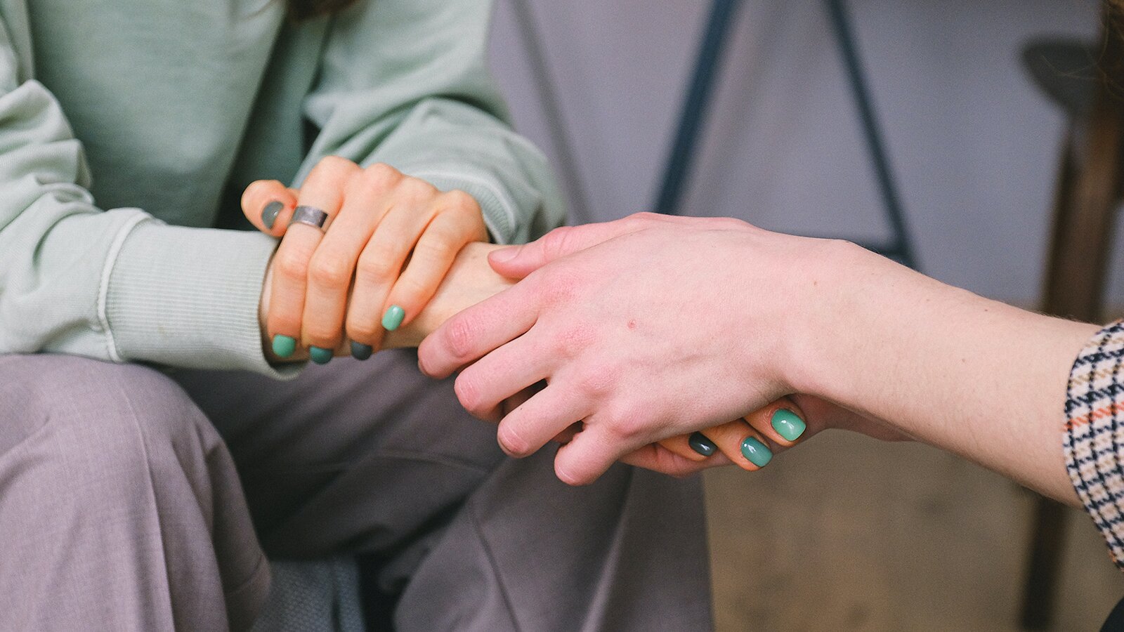 Zdjęcie przedstawia splecione ręce. Po lewej stronie widoczne są ręce oraz fragment jasnozielonej bluzy. Ręce oparte są na kolanach w szarych spodniach. Dłonie mają pomalowane na zielono paznokcie. Lewa dłoń położona jest na nadgarstku prawej ręki, która jest wyprostowana i przytrzymywana w dłoniach postaci po prawej stronie.  