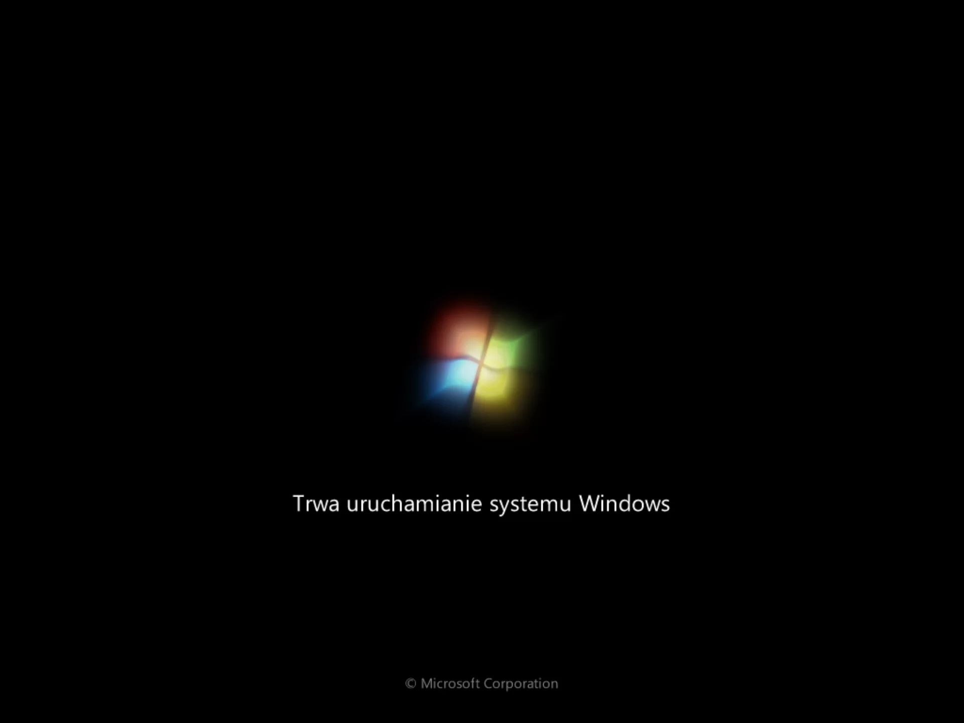 Na zdjęciu pokazano zrzut ekranu powitalnego Windows.W centralnej części zdjęcia na czarnym tle znajduje się logo Windows. Logo jest w kształcie nieznacznie pochylonego okna zbudowanego z czterech falujących kwadratów w kolorach: czerwony, zielony, niebieski, żółty. Poniżej loga znajduje się napis: Trwa uruchamianie systemu Windows.