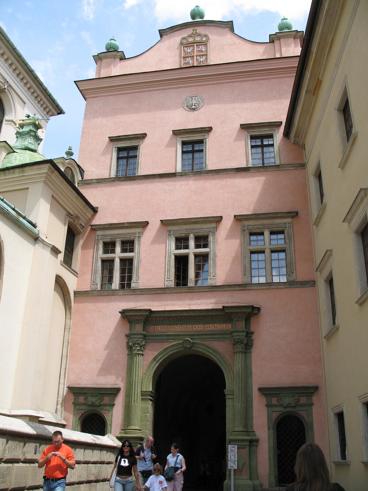 Ilustracja przedstawia bramę wjazdową do zamku królewskiego na Wawelu. Budynek jest dwupiętrowy z trzema oknami na każdym piętrze, zwieńczony attyką i wjazdem zwieńczonym półokrągłym łukiem. Znajduje się pomiędzy zwartą zabudową.