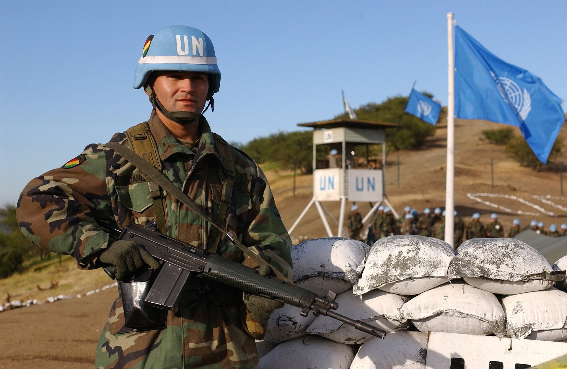 Na zdjęciu żołnierz sił pokojowych ONZ. W ręku trzyma karabin, a na głowie ma niebieski hełm z napisem UN. Stoi obok worków z piasem. W tle flaga ONZ oraz wieżyczka z żołnierzami. W oddali widać wzgórze.