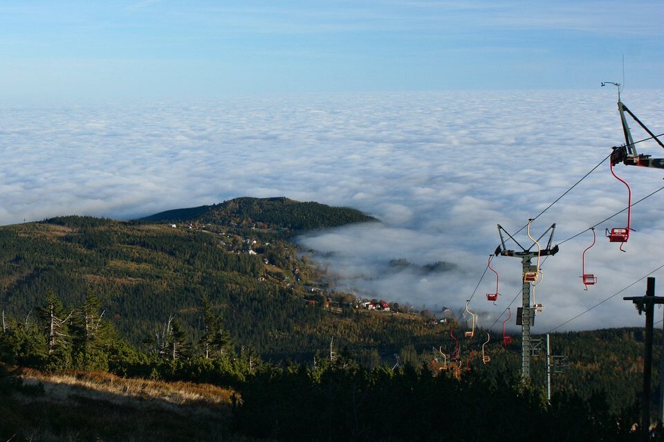 Fotografia barwna. Ilustracja przestawia widok na dolinę, która otaczają chmury. W prawym rogu fotografii znajduje się wyciąg krzesełkowy.