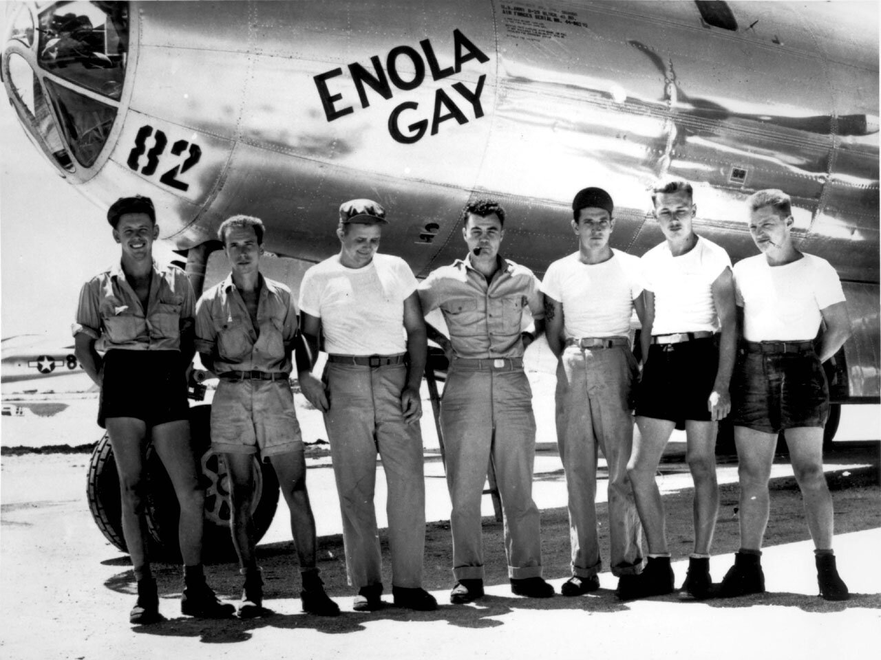 Siedmiu  mężczyzn pozuje do zdjęcia na tle kadłuba samolotu. Większość z nich ubranych jest w letnie stroje - podkoszulki, krótkie spodnie. Stoją w równym szeregu, w swobodnych pozach. Na kadłubie samolotu widnieje napis: ENOLA GAY i numer 82.