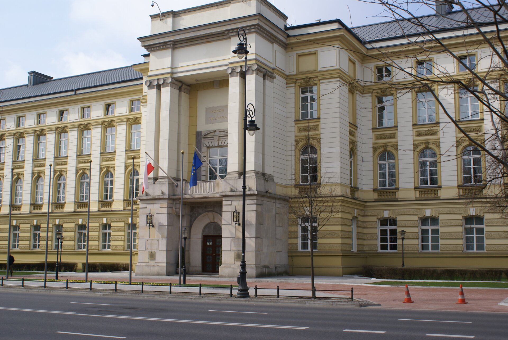 Zdjęcie przedstawia trzypiętrowy budynek. Znajduje się on przy ulicy. Posiada okazałe wejście. Nad wejściem powiewają dwie flagi: Polski i Unii Europejskiej. 