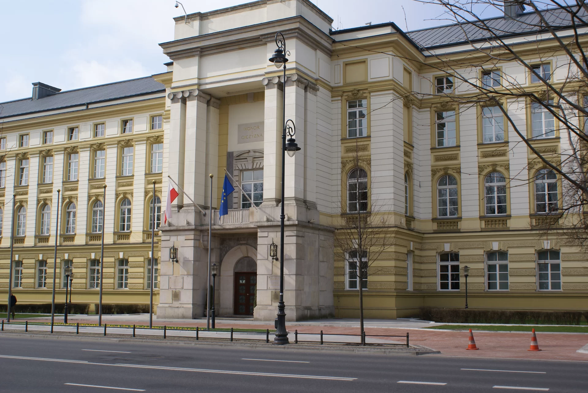 Kliknij, aby powiększyć. Zdjęcie przedstawia trzypiętrowy budynek. Znajduje się on przy ulicy. Posiada okazałe wejście. Nad wejściem powiewają dwie flagi: Polski i Unii Europejskiej.