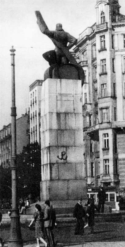 Kliknij, aby powiększyć. Czarno‑białe zdjęcie przedstawia pomnik poświęcony pamięci polskich lotników. Na wysokim postumencie znajduje się rzeźba pilota, który wspiera się o śmigło. Sylwetka widoczna jest od tyłu. W połowie wysokości postumentu widnieje namalowana czarną kotwicą (symbolem Polski Walczącej). Przed pomnikiem widać ludzi - mężczyzna stoi i przygląda się, z boku dwie osoby siedzą na cokole, dwie pary przechodzą. W tle widać budynki i drzewa.