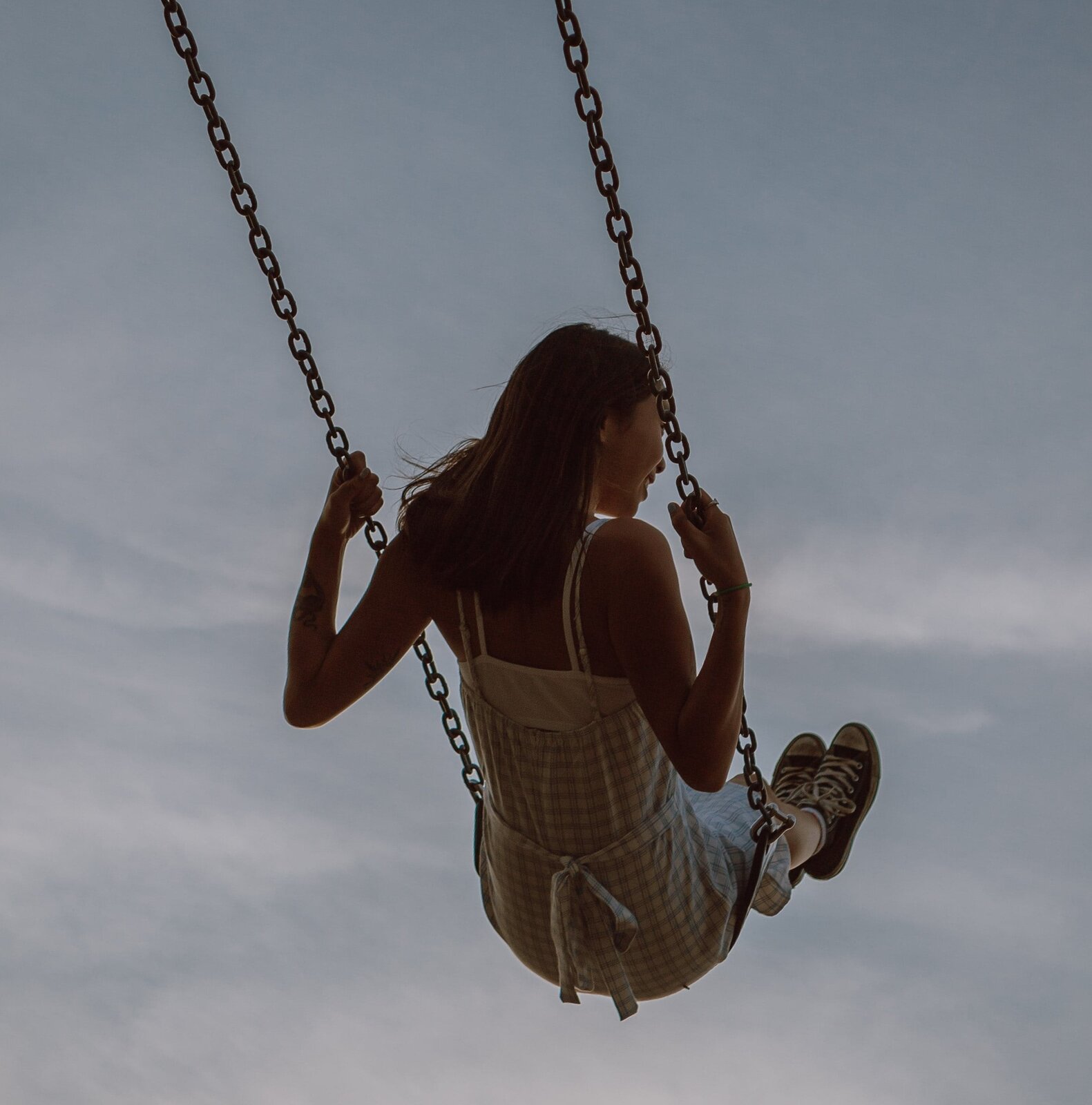 Fotografia przedstawia dziewczynkę na huśtawce. Huśtawka składa się z łańcucha i skórzanego pasa służącego za siedzisko. Długie włosy dziewczynki są rozwiane, w tle pokryte chmurami niebo.