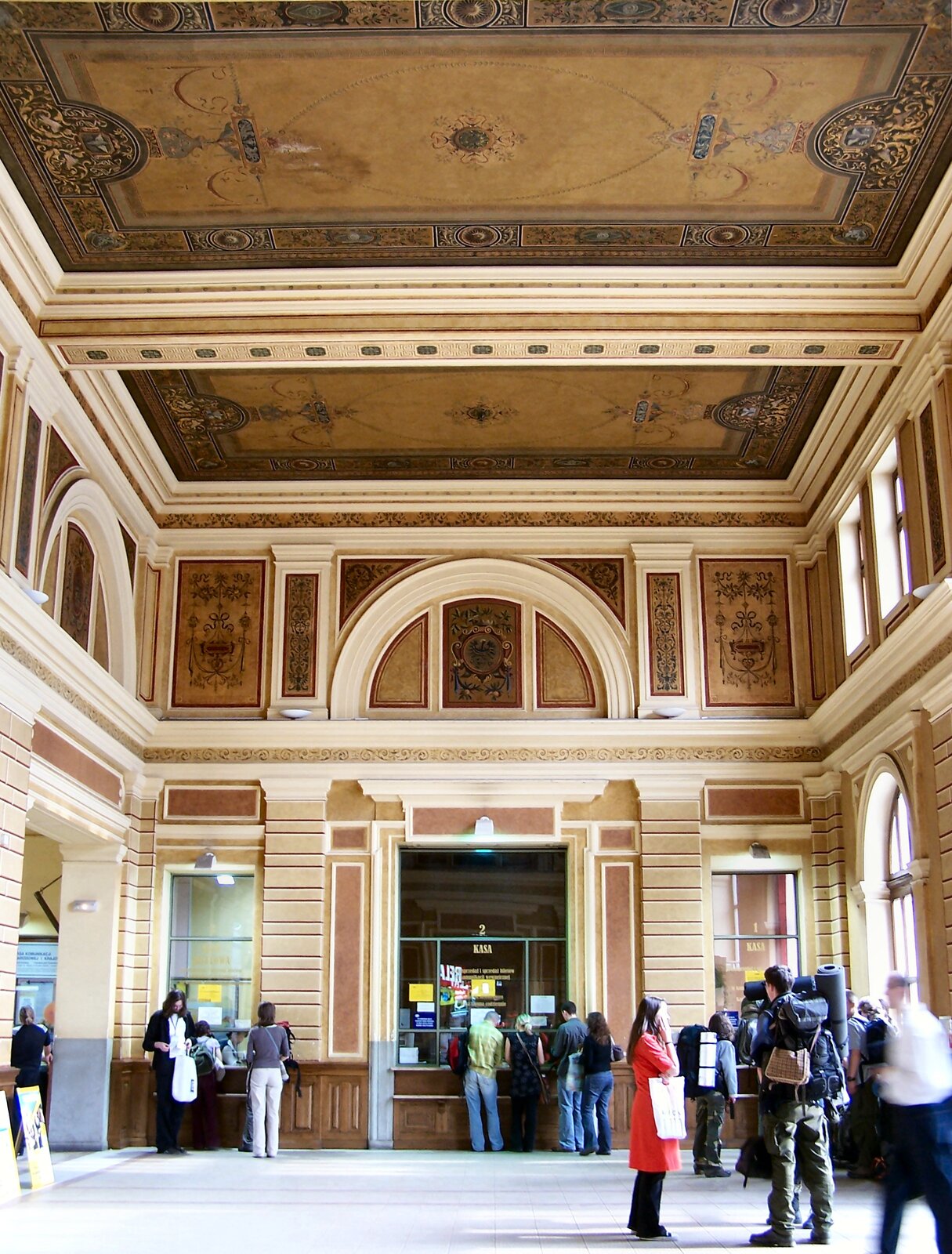 Zdjęcie przedstawia fragment hali dworcowej. Sufit ozdobiony pięknymi freskami. Przy okienku kas biletowych stoją  pasażerowie kupujący bilety. 