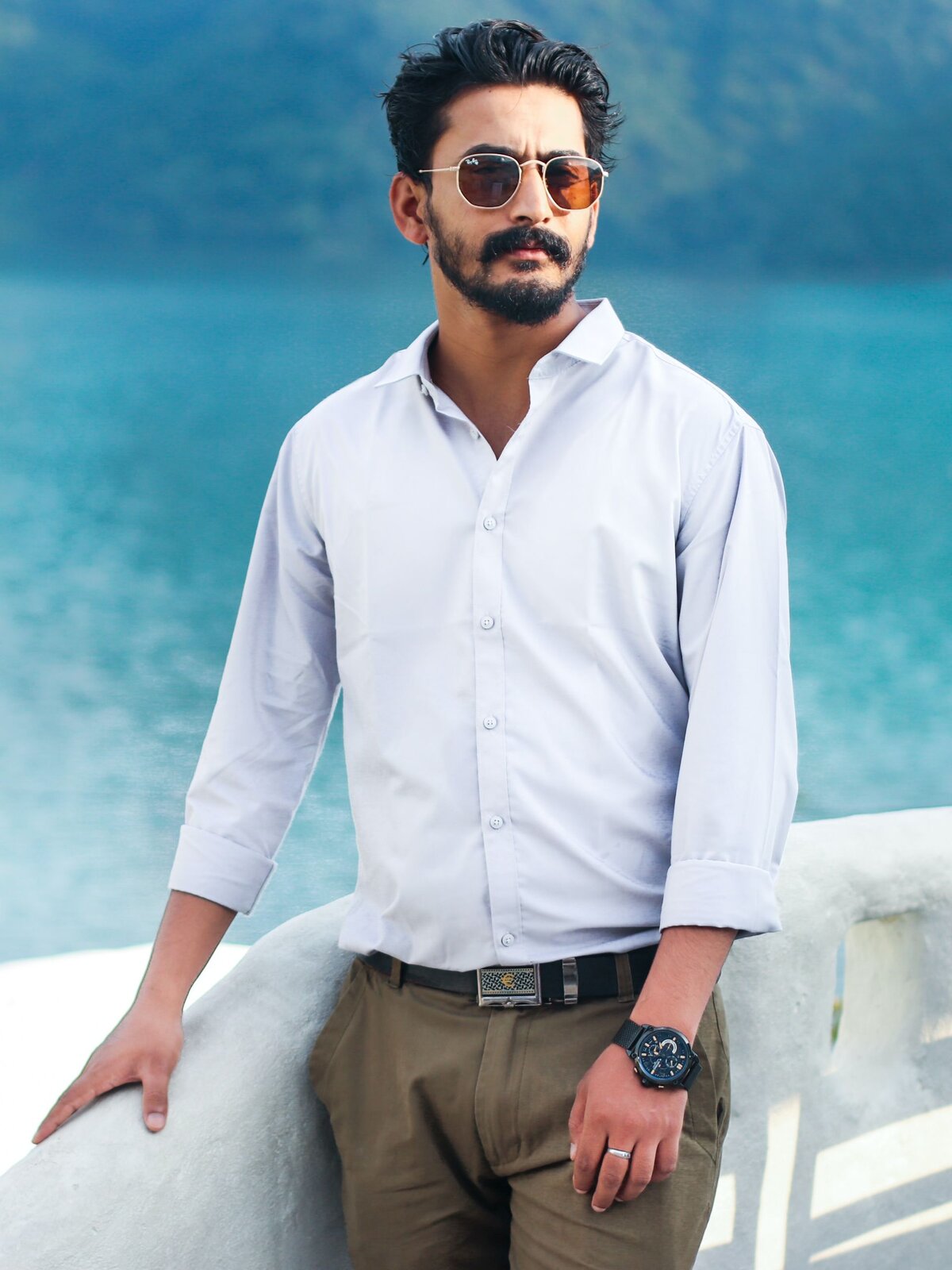 Zdjęcie przedstawia młodego mężczyznę ubranego w białą koszulę rozpiętą pod szyją oraz spodnie w kolorze jasnego brązu z paskiem mającym ozdobną klamrę. Na twarzy widoczne okulary przeciwsłoneczne. 