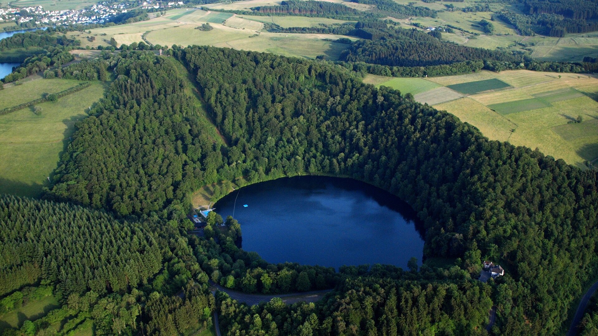Zdjęcie przedstawia maar. Jest to zagłębienie terenu wypełnione wodą, jego zbocza porastają drzewa. W tle znajdują się pola.