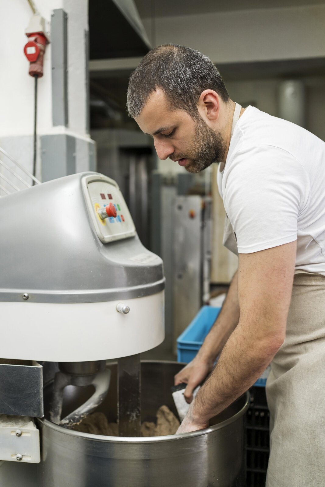 Zdjęcie drugie, podpisane Użytkowanie urządzeń do produkcji pieczywa. Na zdjęciu widać mężczyznę w beżowym fartuchu. Obsługuje duży robot do produkcji pieczywa. Wewnątrz ogromnego garnka podstawionego pod maszynę widoczne jest surowe ciasto.