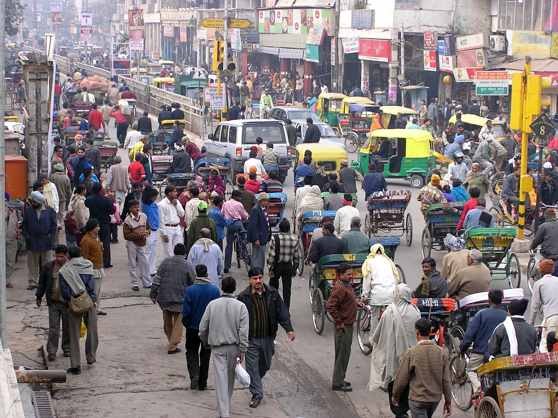 Na zdjęciu widoczna jest ulica w Delhi. Panuje duży tłok. Na chodnikach  i na ulicy tłoczą się przechodnie, samochody, riksze. Po prawej stronie ulicy widać gęstą zabudowę punktów usługowych i domów oblepionych reklamami i szyldami. 