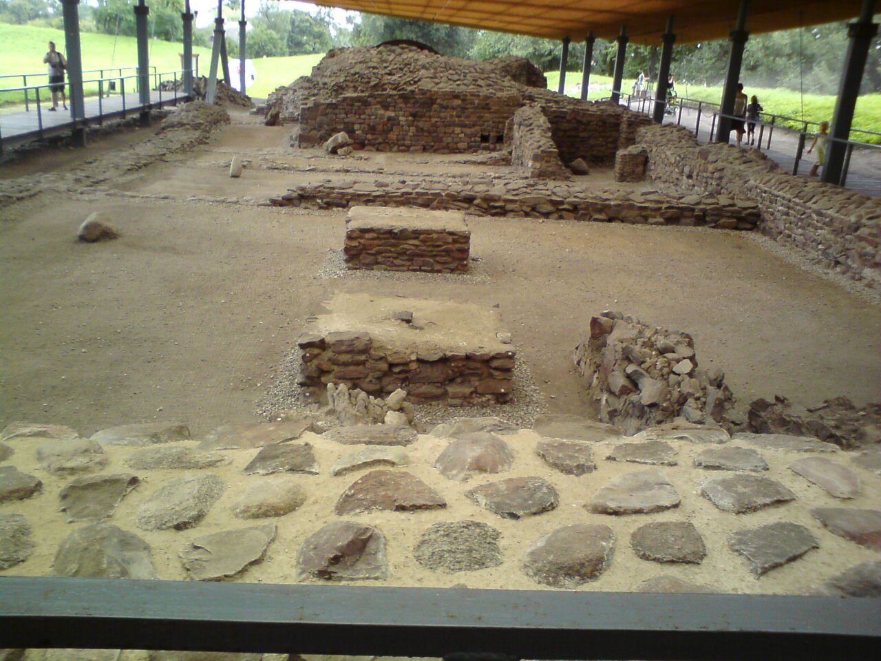 Zdjęcie przedstawia zadaszone ruiny. Zachowały się murki wykonane z kamienia. Dookoła ruin chodzą ludzie.