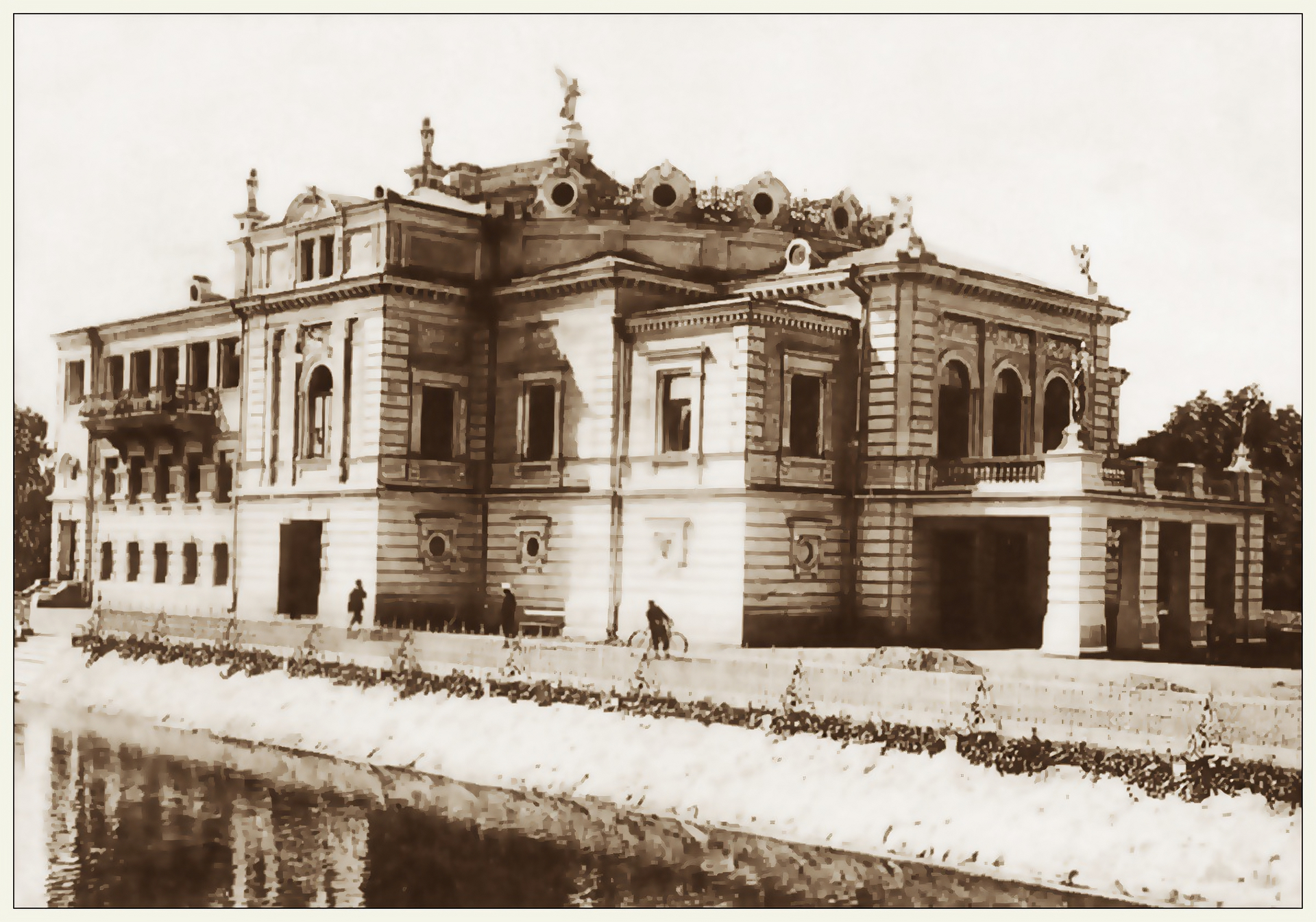 Zdjęcie przedstawia neorenesansowy gmach nowego teatru miejskiego. Budowla jest dwukondygnacyjna w stylu palladiańskim. Wejście zlokalizowane jest pod portykiem kolumnowym, nad którym góruje taras widokowy. W bocznej części budynku, na wyższej kondygnacji znajduje się taras stojący na czterech kolumnach. Przed budynkiem przechadzają się ludzie. Budynek stoi nad rzeką.