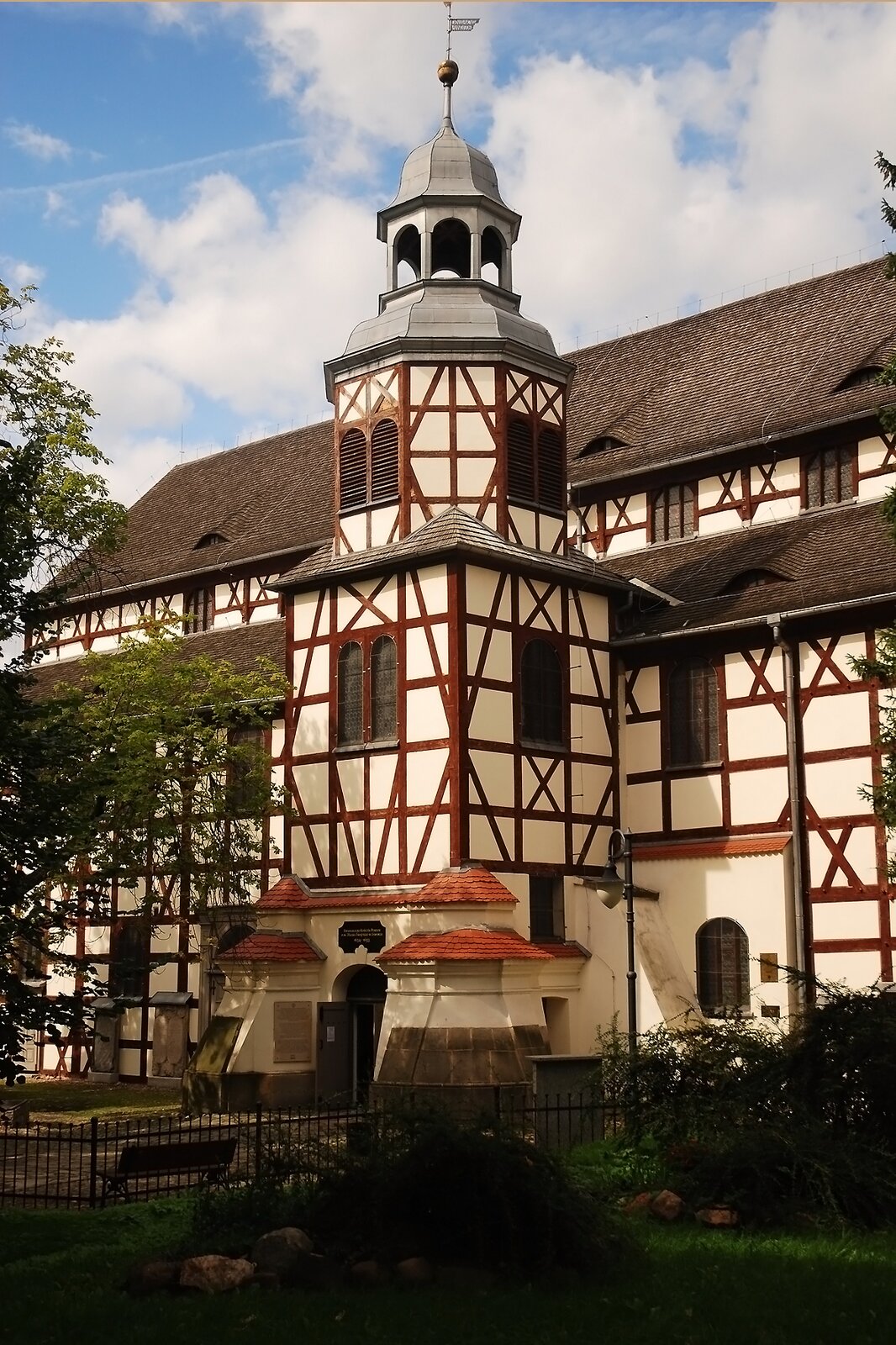 Zdjęcie przedstawia kościół pokoju w Jaworze. Na pierwszym planie na środku jest kawałek placu z ławkami i bramą. Po obu stronach są wysokie drzewa. Dalej jest kościół. Wejście znajduje się w dobudowanej wieży. Budynek ma białe ściany poprzecinane brązowymi drewnianymi deskami. Okna posiadają okiennice.