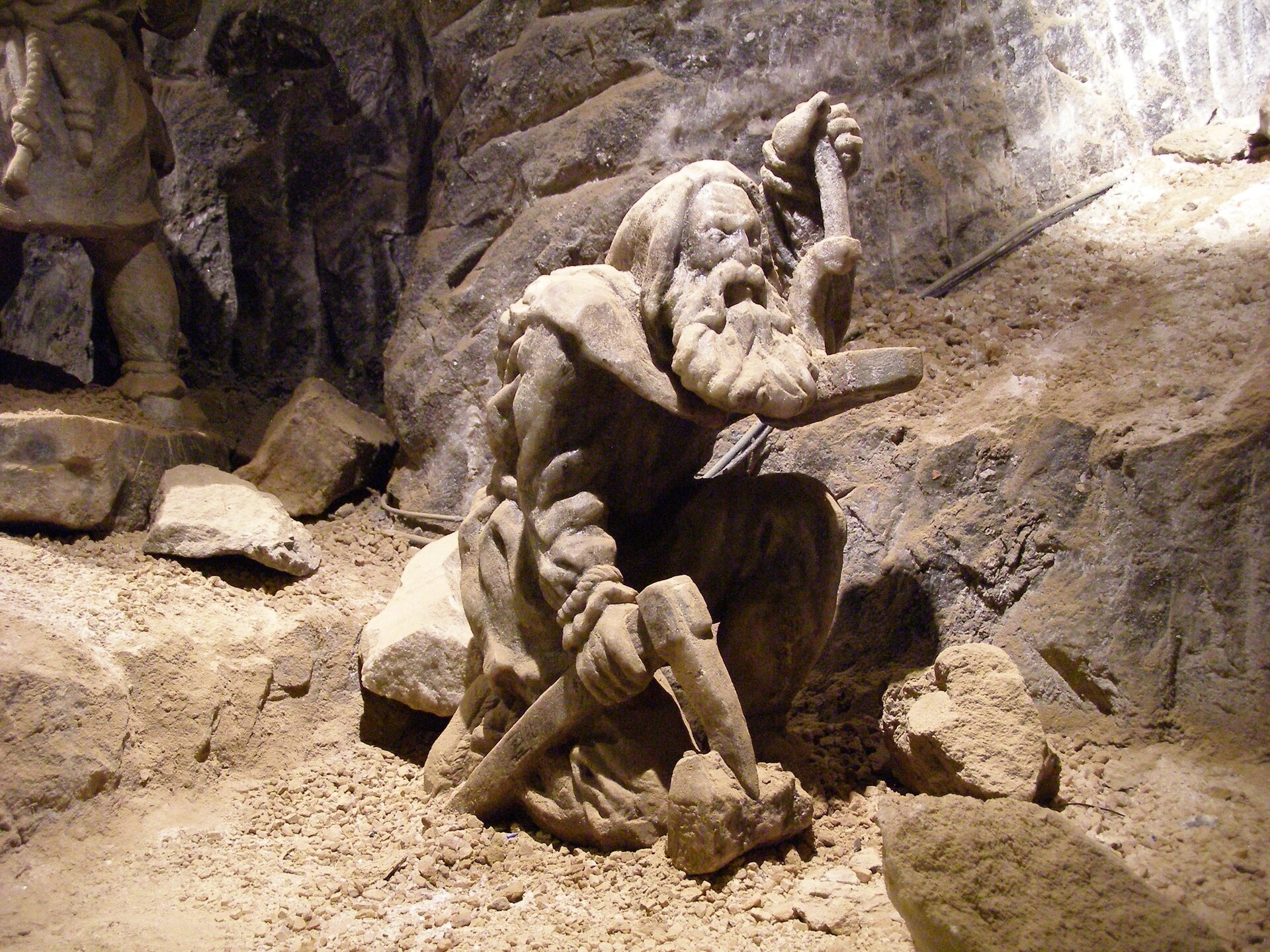 Zdjęcie ukazuje rzeźbę solną - kruszaka z kagankiem i kilofem. Mężczyzna siedzi na kamieniu. Ma długą brodę. W jeden dłoni trzyma kaganek, w drugiej kilof na kamieniu. Wokół rzeźby są kawałki skał i skalna ściana.