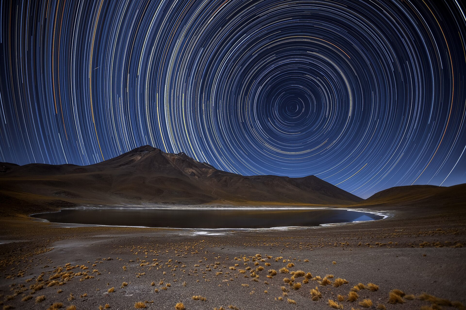 Fotografia przedstawia pustynny krajobraz nocą. Pośrodku, wśród porośniętej gdzieniegdzie żółtymi, niskimi kępkami roślin gleby, widać owalny zbiornik wodny, odbijający kolor zbocza niewysokich gór widocznych tuż za nim. Gleba oraz góry są w odcieniu szarobrązowym, z widocznymi gdzieniegdzie żółtymi roślinami. Góry oddzielają fragment ziemi od nieba, którego ciemnoniebieski odcień jest ozdobiony kolorowymi pasmami. Po prawej stronie, niedaleko centrum fotografii, widoczny jest mały pierścień złożony z fragmentów niebieskich oraz żółtych zaokrąglonych linii. Następnie linie stają się coraz większe, tworząc pierścienie otaczające pierwszy z nich. Rozciągają się one na całej widocznej powierzchni nieba, komponując mozaikę. 