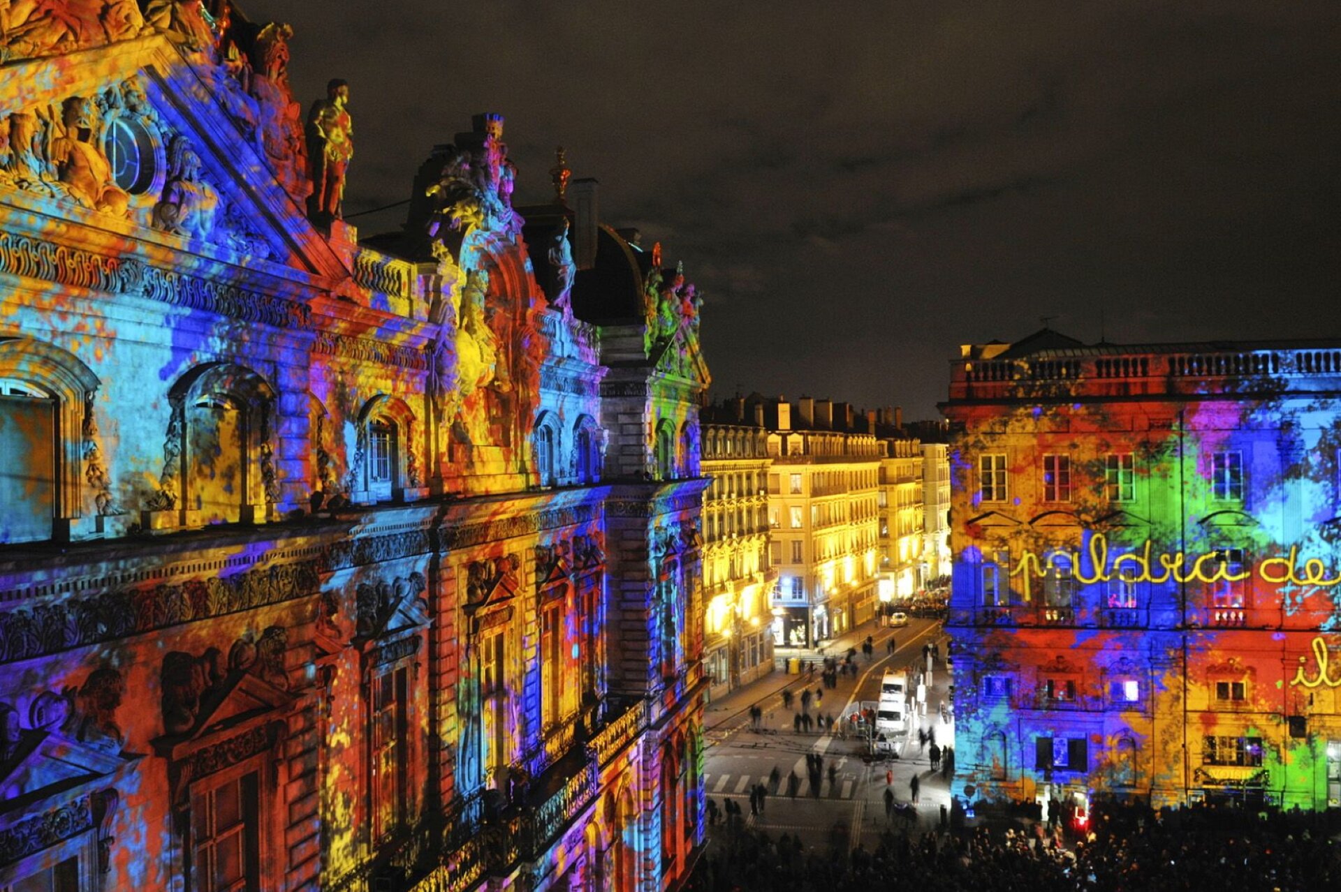 Na zdjęciu znajduje się budynek, na którym wyświetlony został kolorowy obraz. Na elewacji budynku wyświetlone zostały następujące barwy: żółty, pomarańczowy, czerwony, błękitny, niebieski, fioletowy, zielony. Na placu obok budynku stoi tłum ludzi. W tle znajduje się oświetlona ulica, beżowe kamienice i ciemne, nocne zachmurzone niebo. 