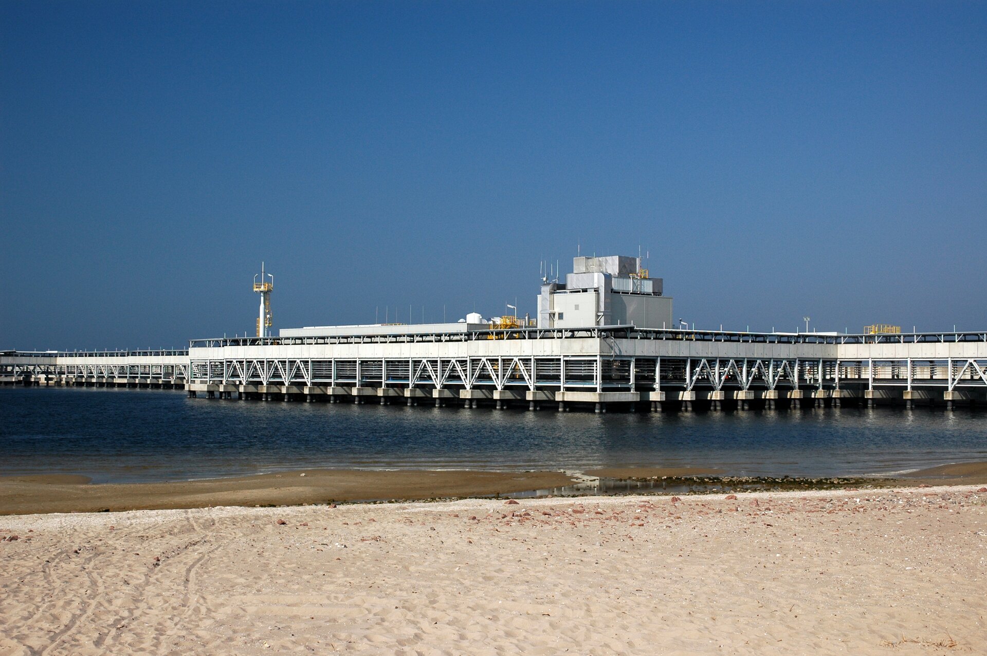 Zdjęcie przedstawia nabrzeże w Świnoujściu. Na pierwszym planie jest piasek, dalej morze oraz zbudowana na palach biała konstrukcja służąca do przeładunku LNG.