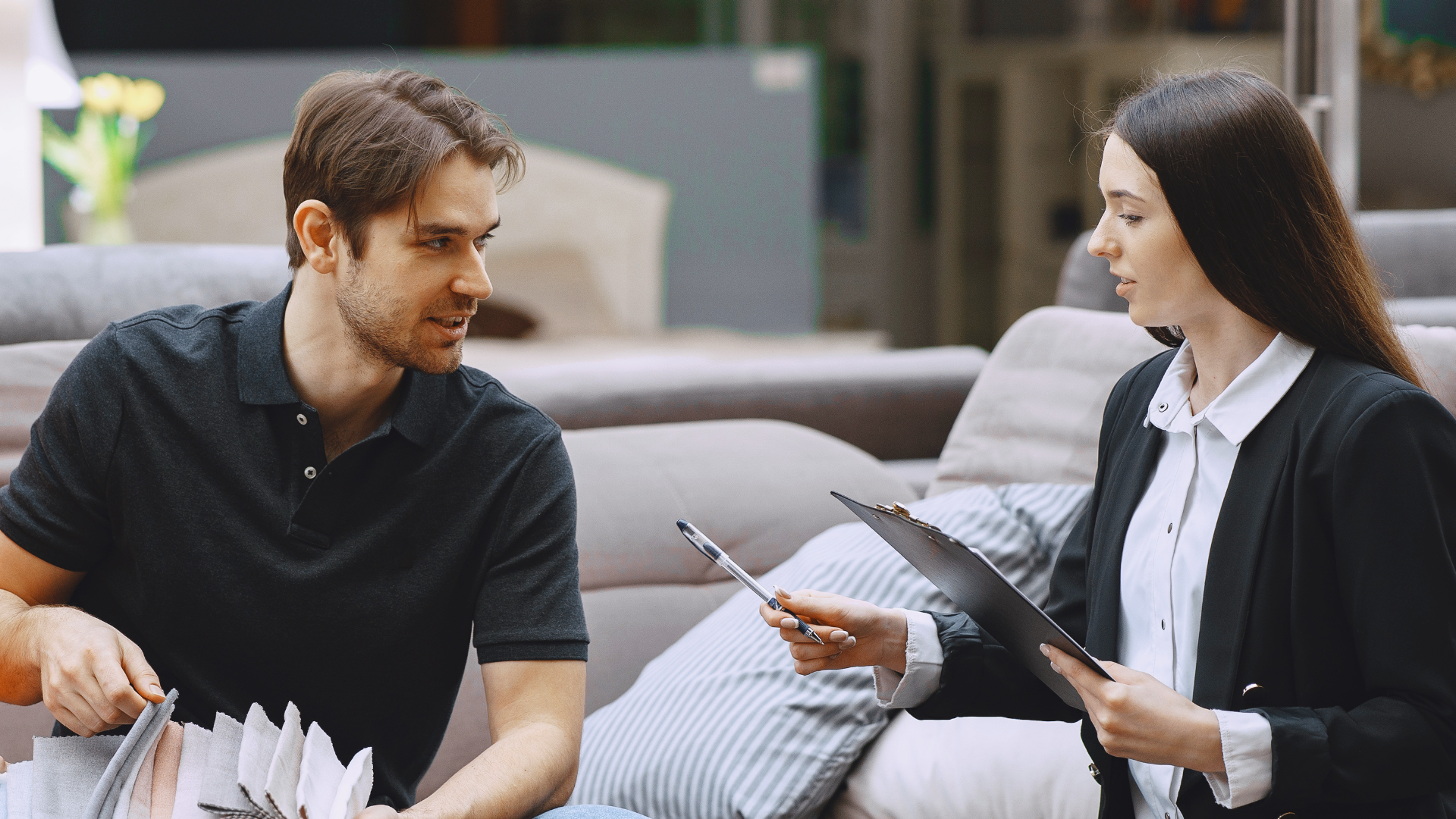 Zdjęcie przedstawia mężczyznę i kobietę w sklepie meblowym na tle tapicerowanych mebli. Po lewej stronie znajduje się mężczyzna – klient, oglądający próbki materiałów obiciowych.  Po prawej stronie znajduje się kobieta – sprzedawczyni. W rękach trzyma notatnik i długopis.  